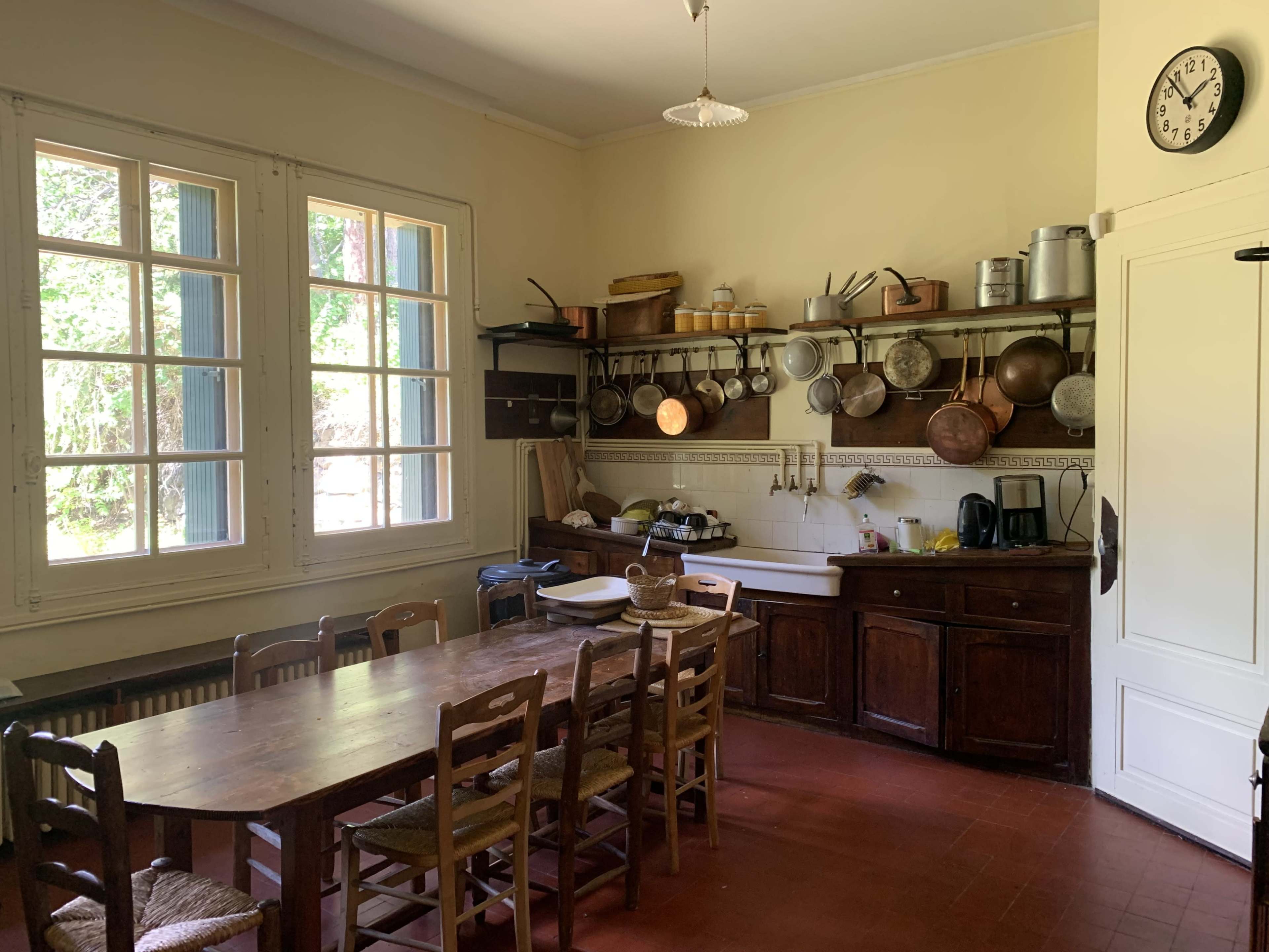 The image shows a spacious kitchen with a wooden dining table, multiple chairs, and various cooking utensils hanging on shelves against the wall.