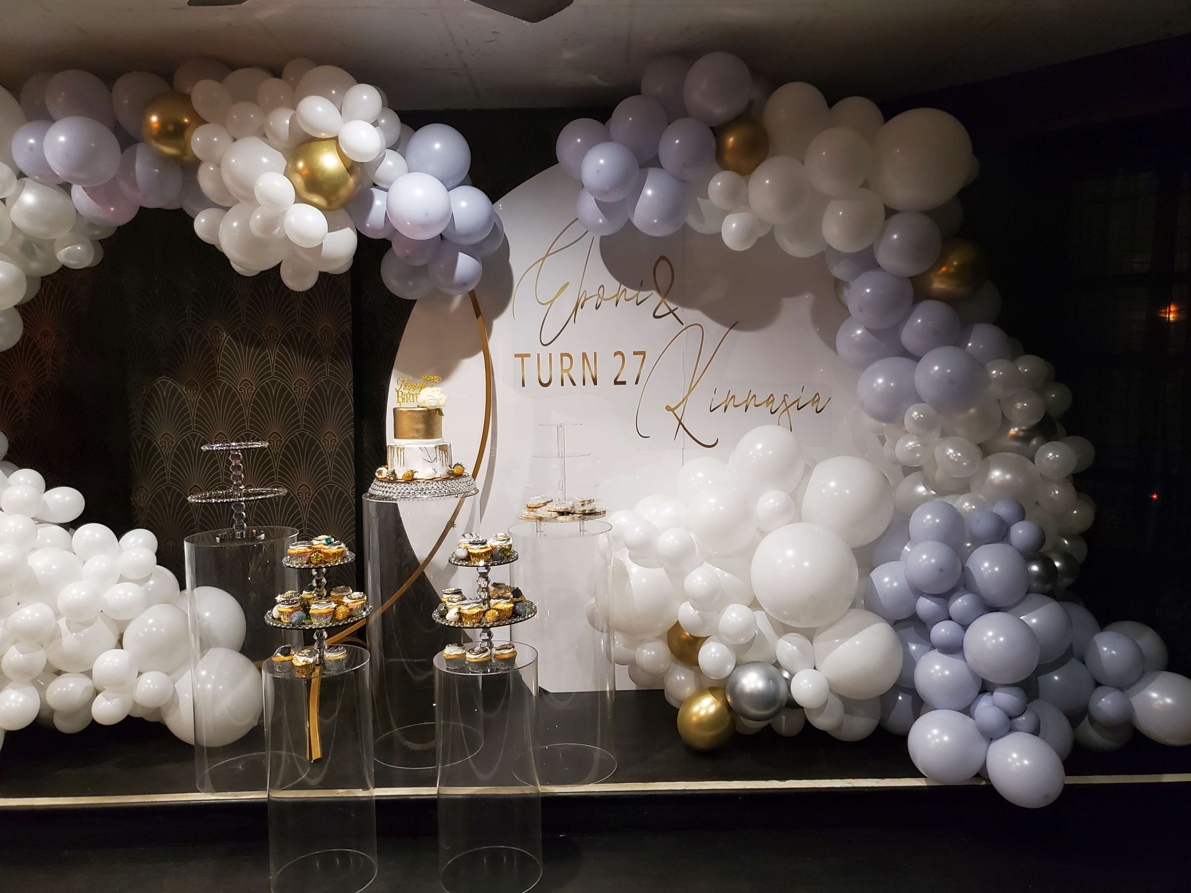 A decorated event space with a large balloon arch in shades of white, gray, and gold, accompanied by a cake and dessert display on clear stands.