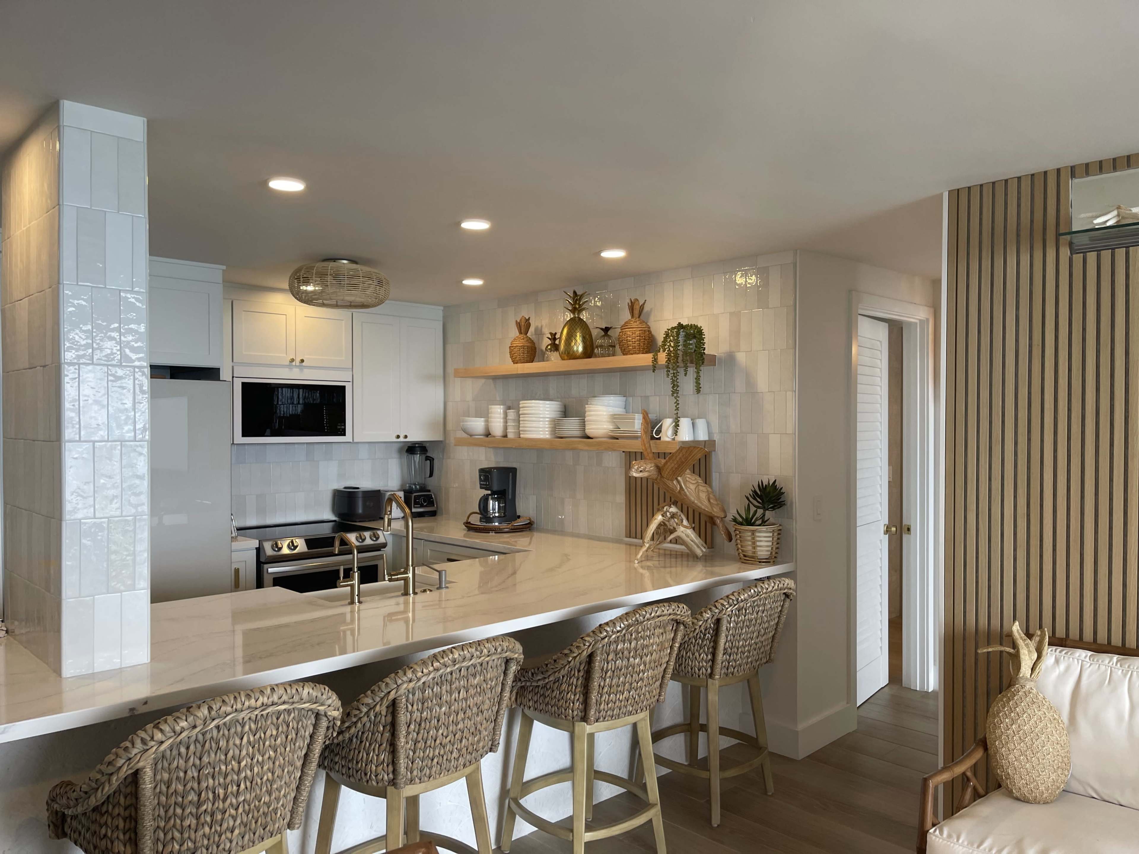 A modern kitchen with white cabinets, a marble countertop, and bar stools, featuring open shelving that displays dishware and decorative plants.