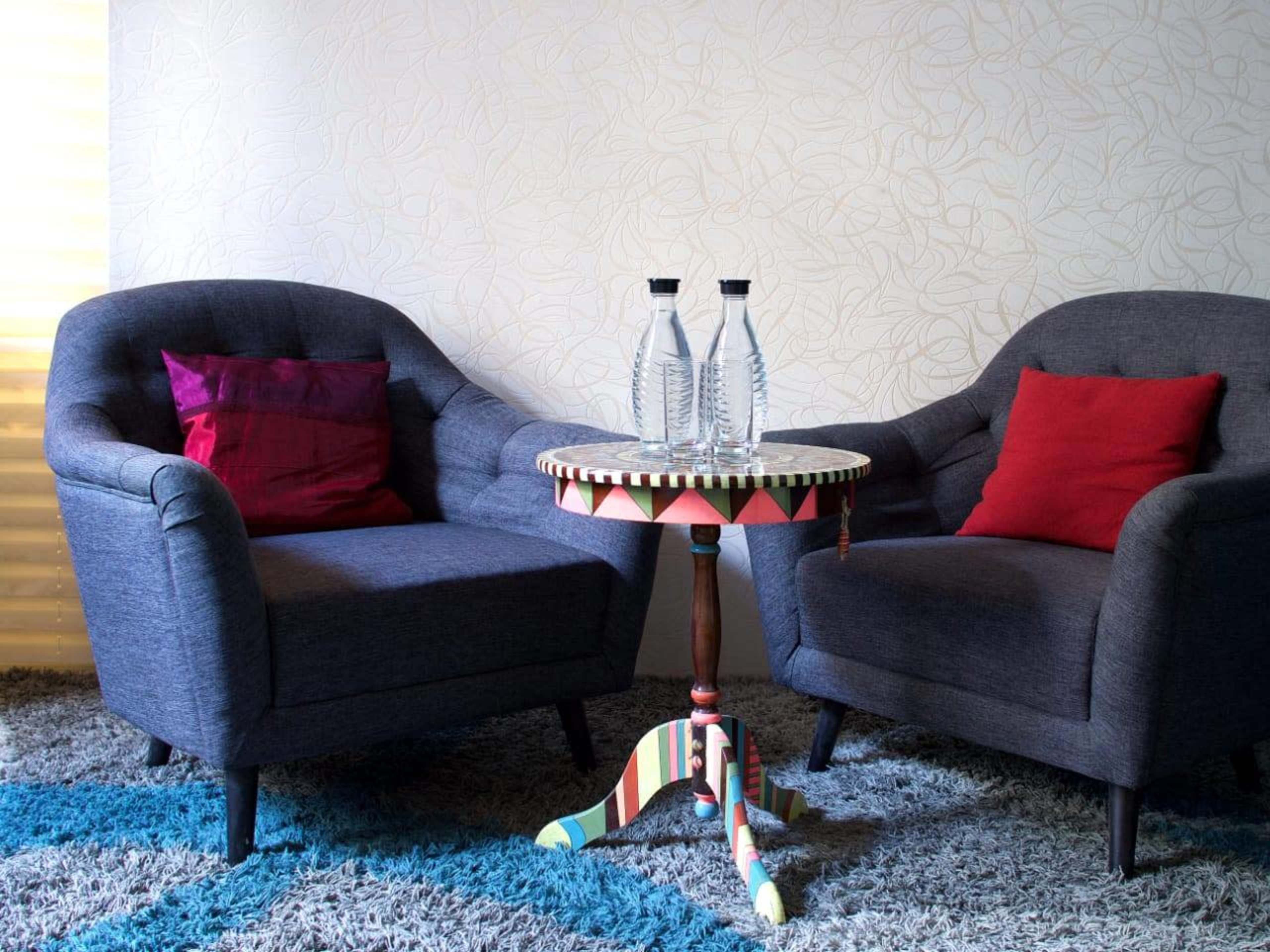 Two gray armchairs with red pillows on either side of a circular table, which has three clear bottles on it, set on a textured rug.