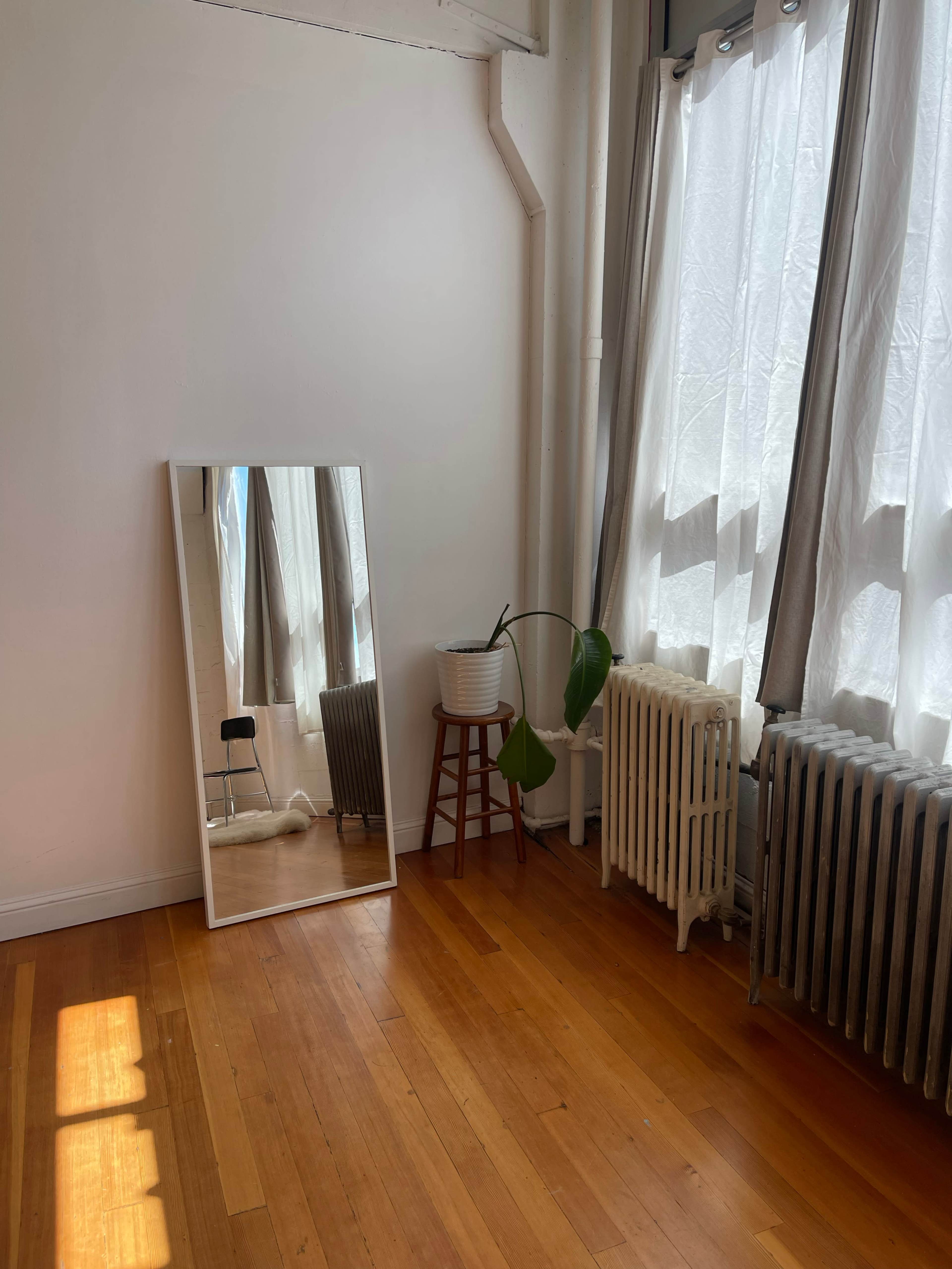 A minimalist room with a tall mirror, a potted plant on a wooden stool, and two metal radiators beside a window adorned with sheer curtains.