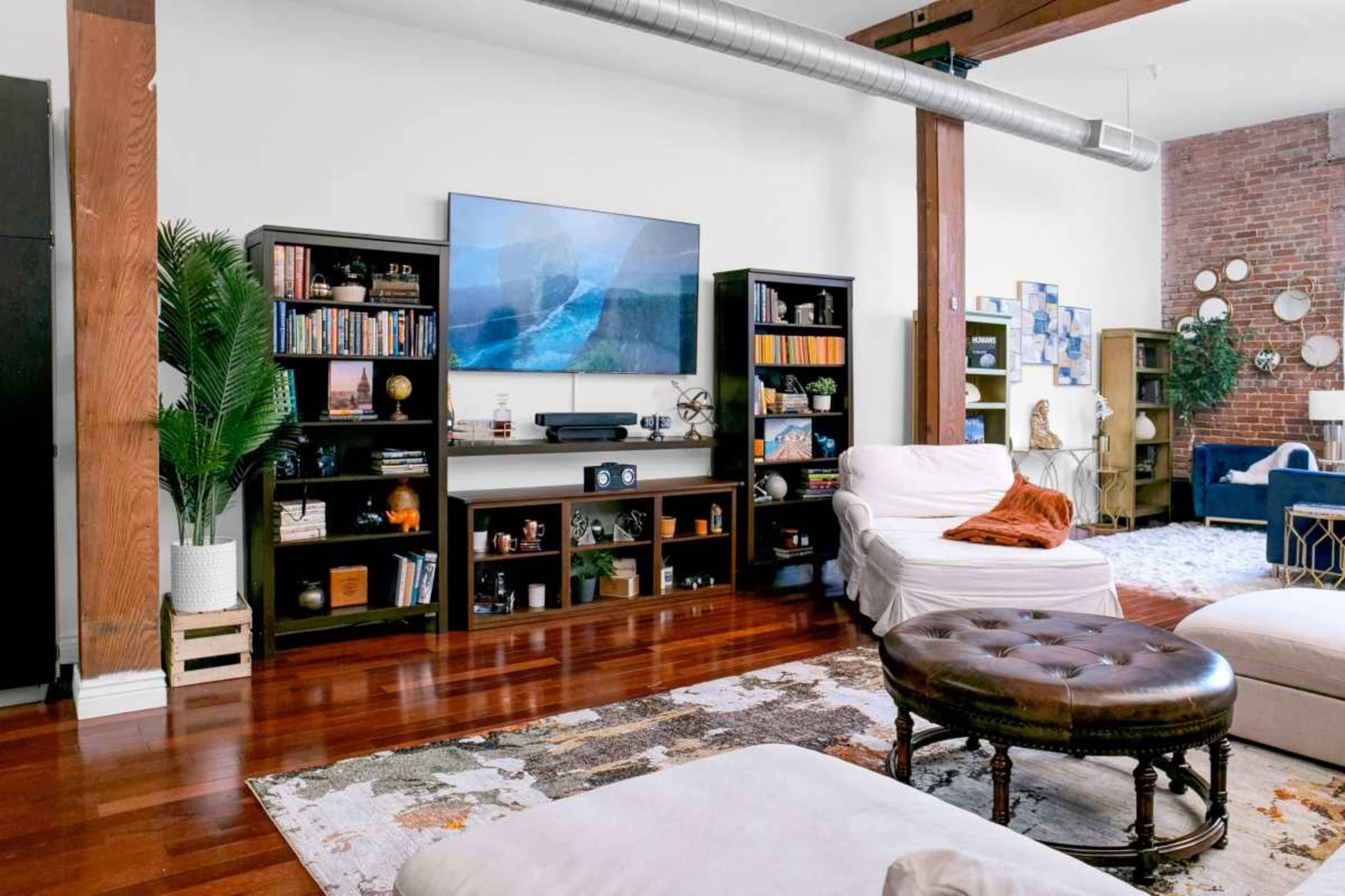 The image shows a modern living room with wooden floors, a large flat-screen TV mounted on the wall, bookshelves filled with books and decor, and a mix of upholstered seating arrangements.
