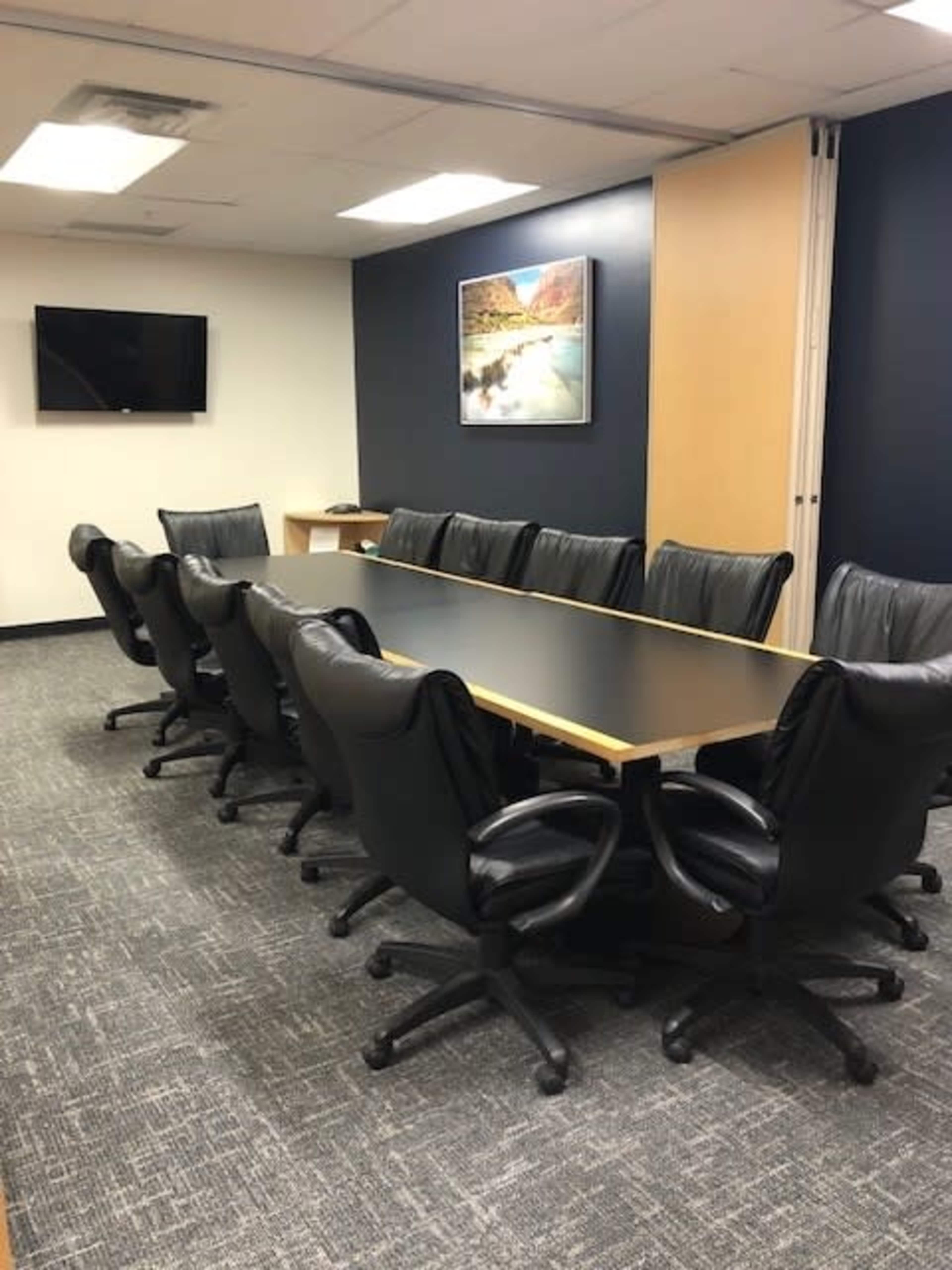 A long conference table with black leather chairs is arranged in a modern office meeting room featuring a wall-mounted television and a landscape picture.