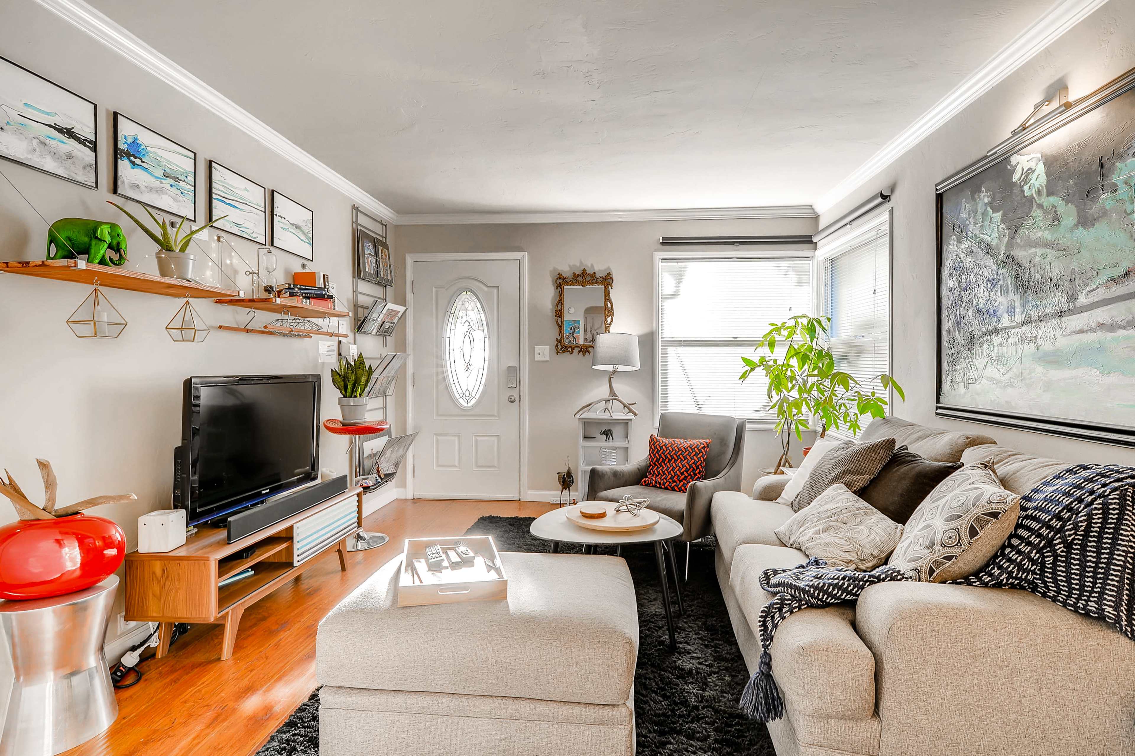 The image shows a cozy living room with a grey sofa, a television on a wooden stand, a round coffee table, and decorative plants along with wall art.