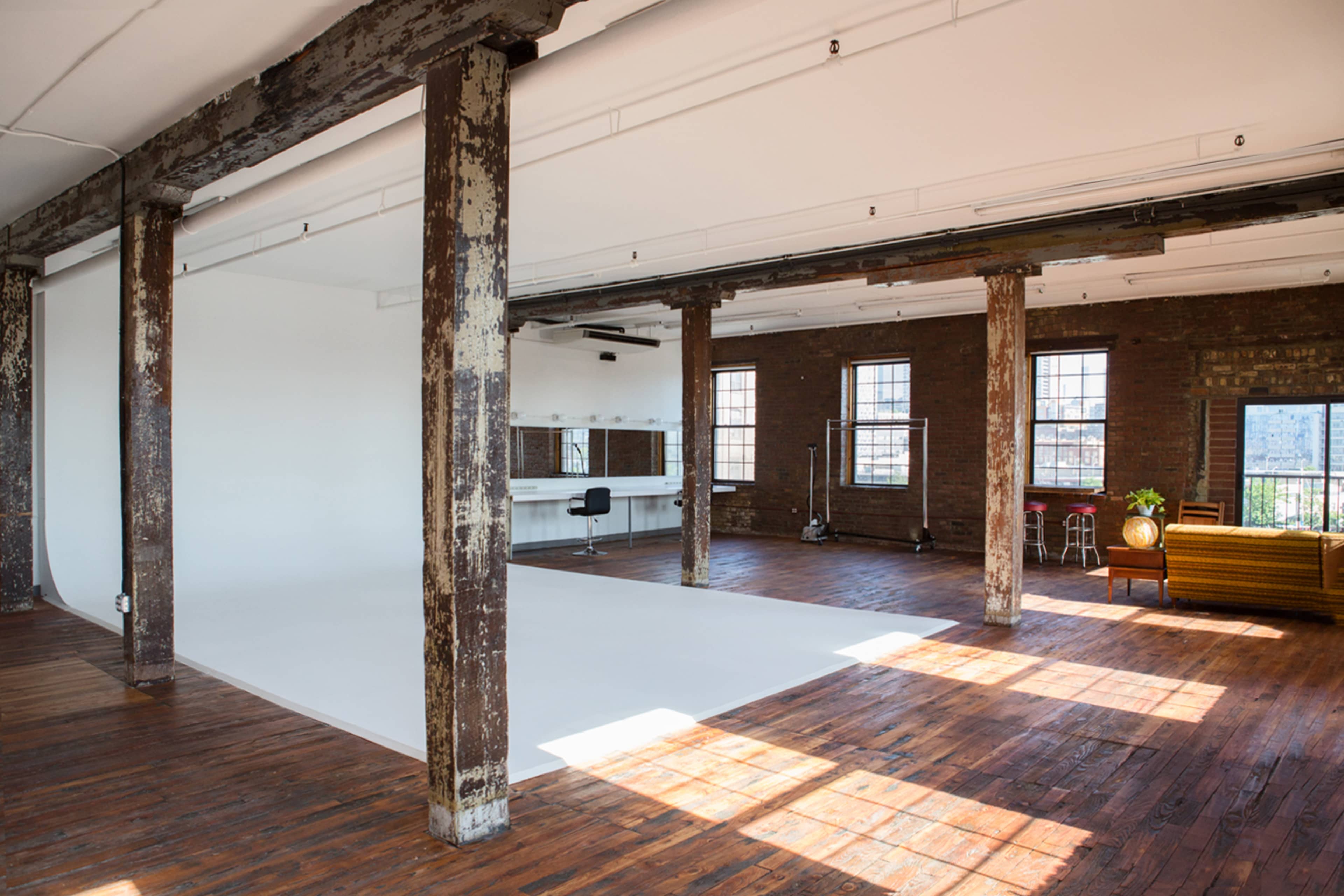 An industrial-style studio features wooden floors, large windows, and exposed brick walls, with a white backdrop and furniture set against the space.