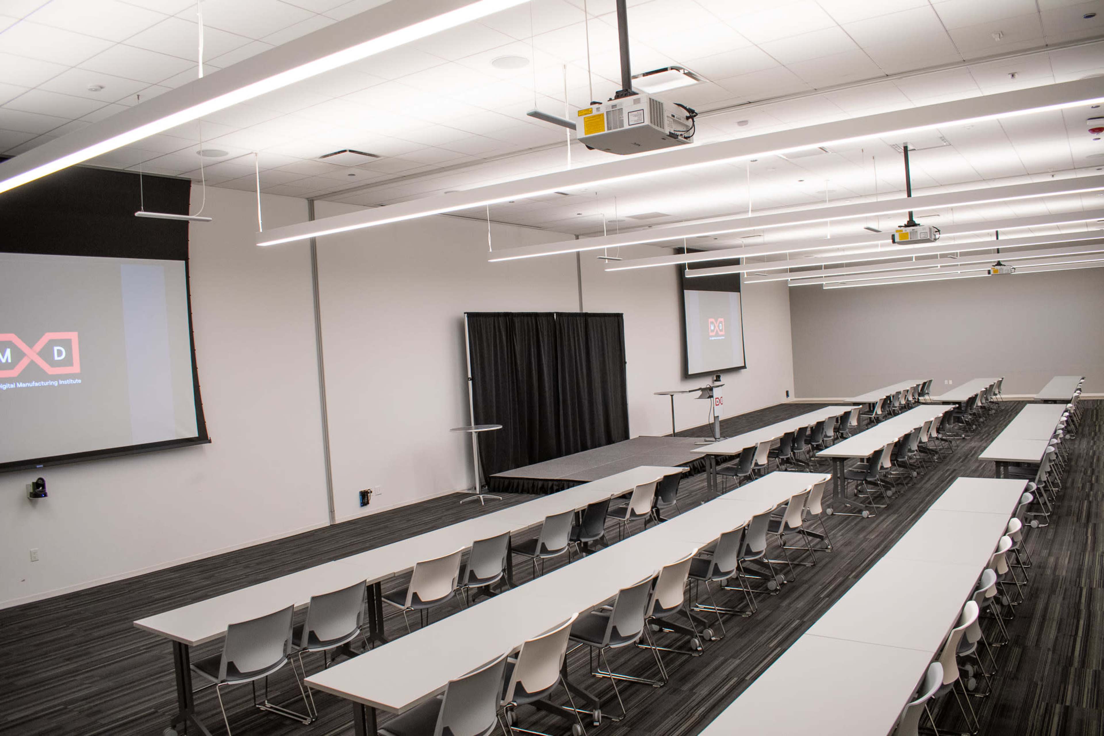 The image shows a spacious conference room set up with multiple long tables and chairs, facing projection screens on opposite walls.