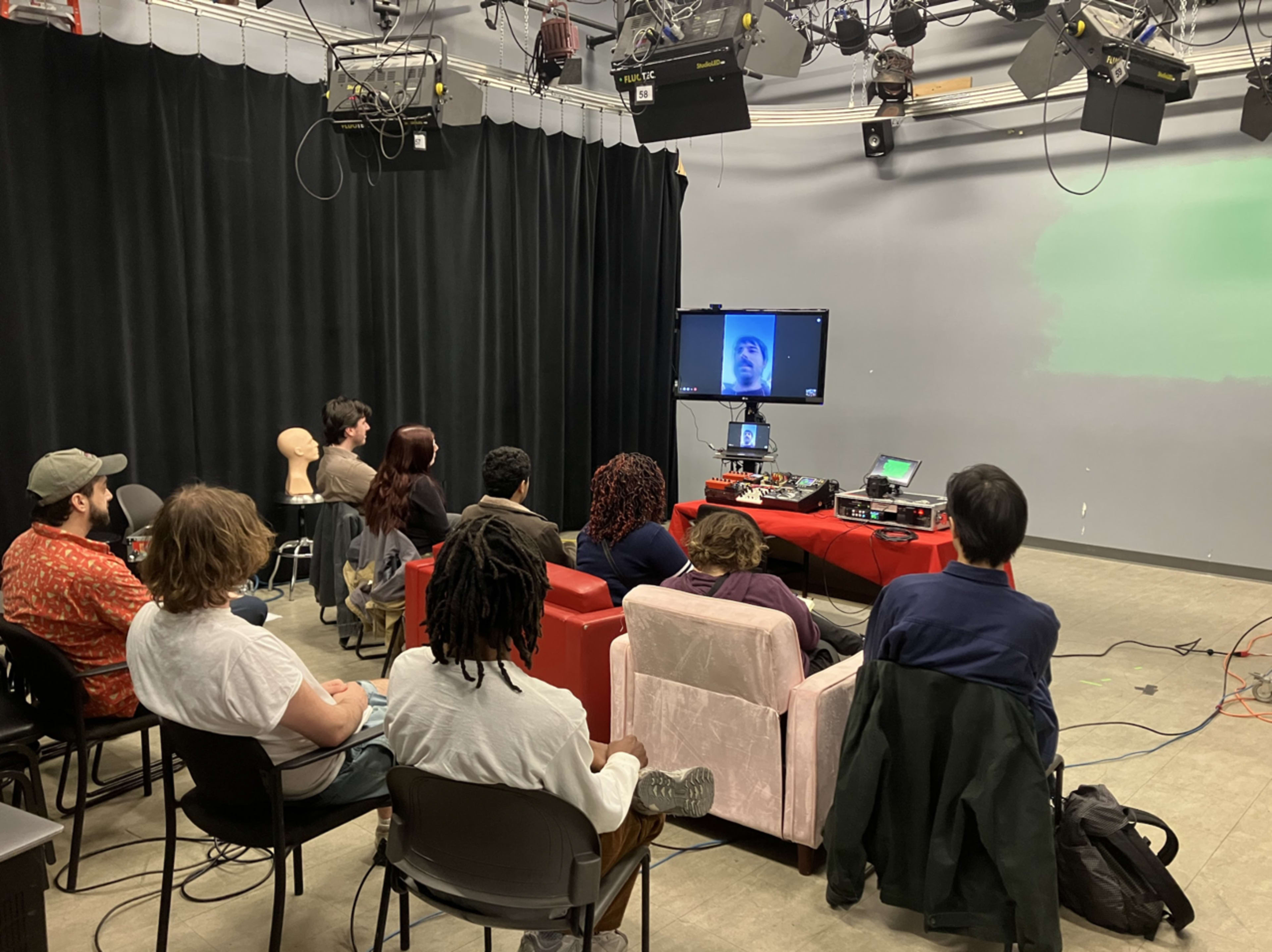 A group of people sits in a studio setting, watching a video conference on a screen in front of them, while various equipment and lighting are visible.