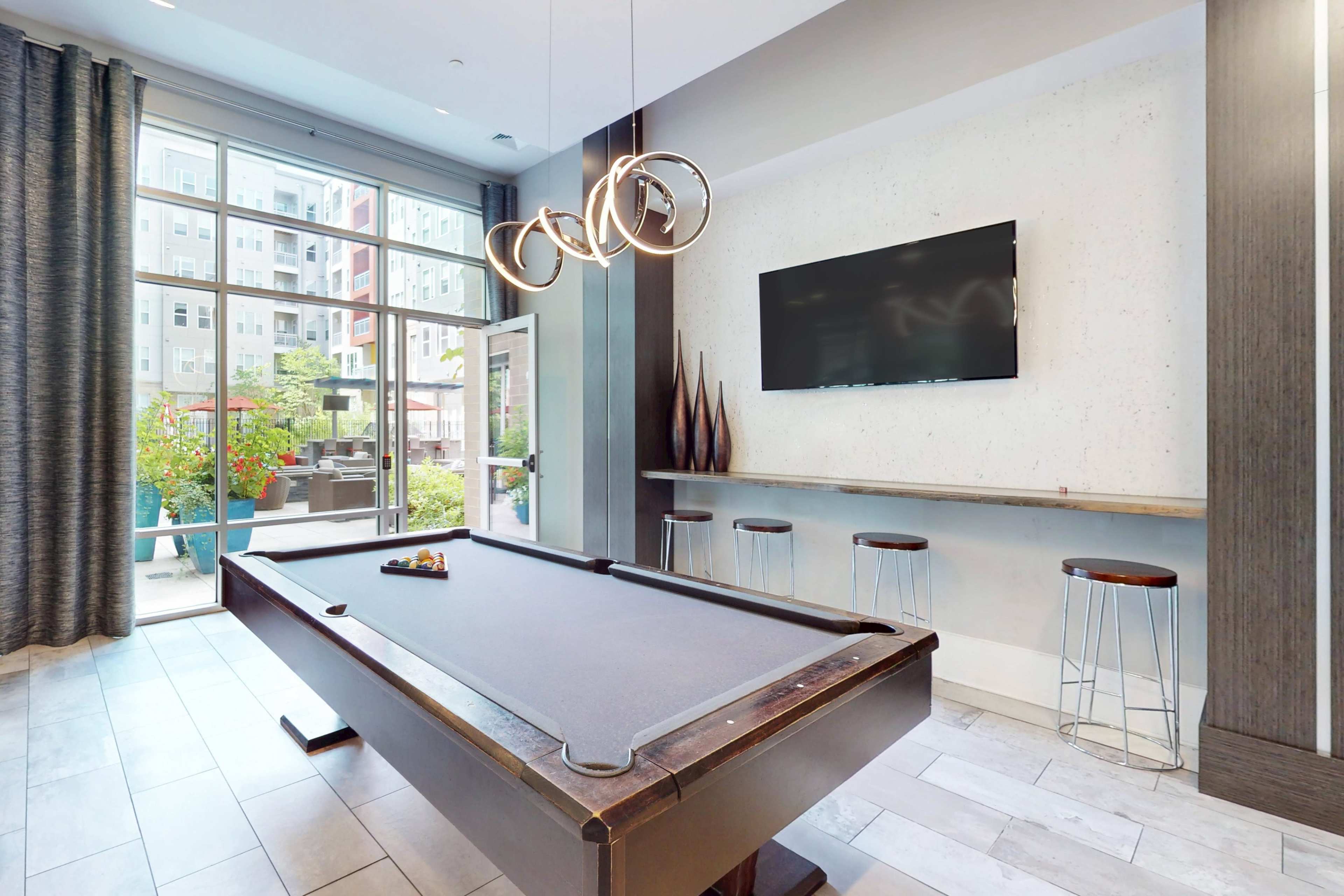 The image features a sleek billiards table in a well-lit lounge area with modern decor, a flat-screen TV mounted on the wall, and bar stools arranged along a counter.