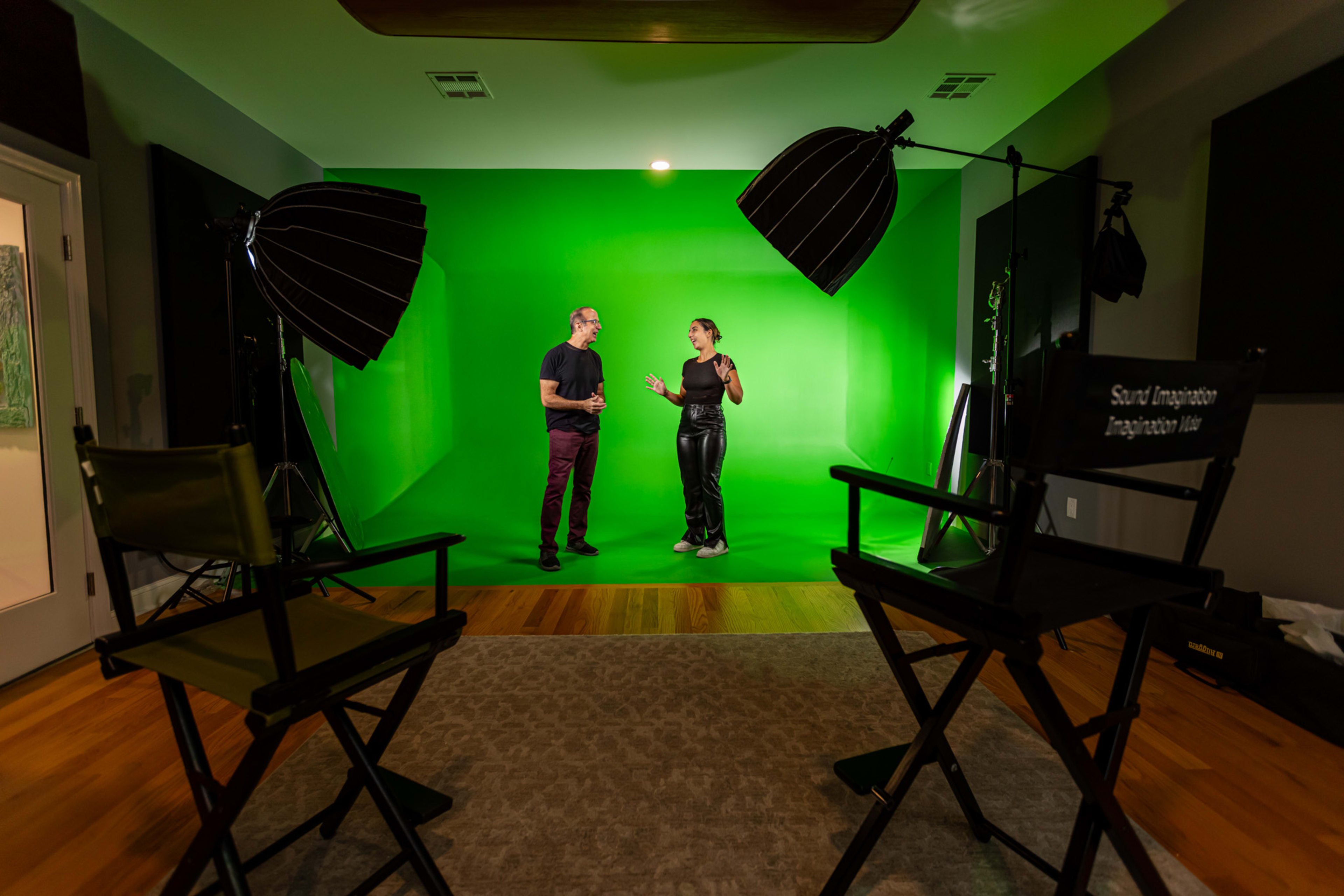 A green screen studio is set up with two people discussing in front of the backdrop, accompanied by studio lights and director's chairs nearby.