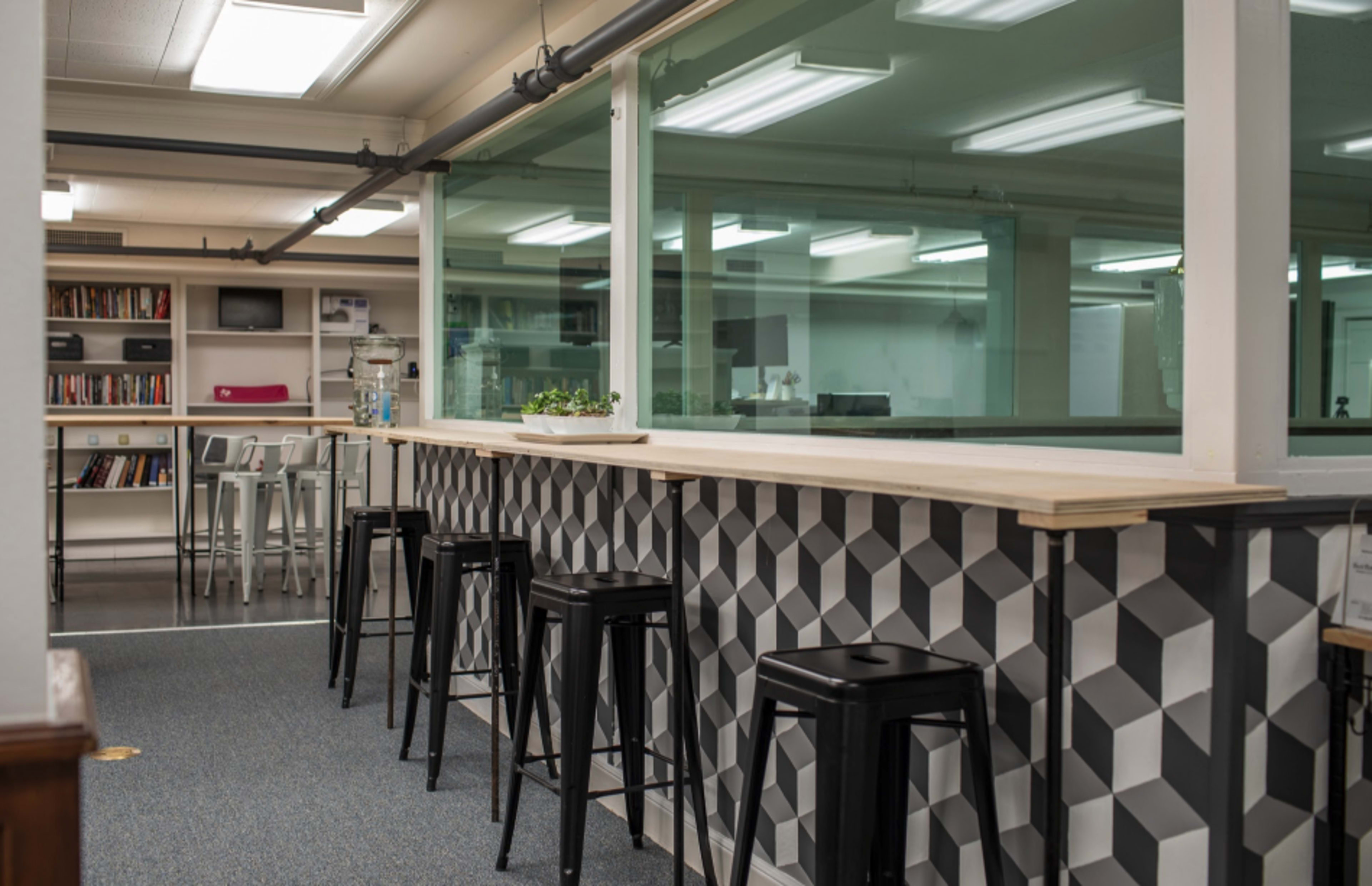 A modern office space features a bar-height table with black stools and geometric patterned panels, overlooking a glass-enclosed area filled with bookshelves and workstations.