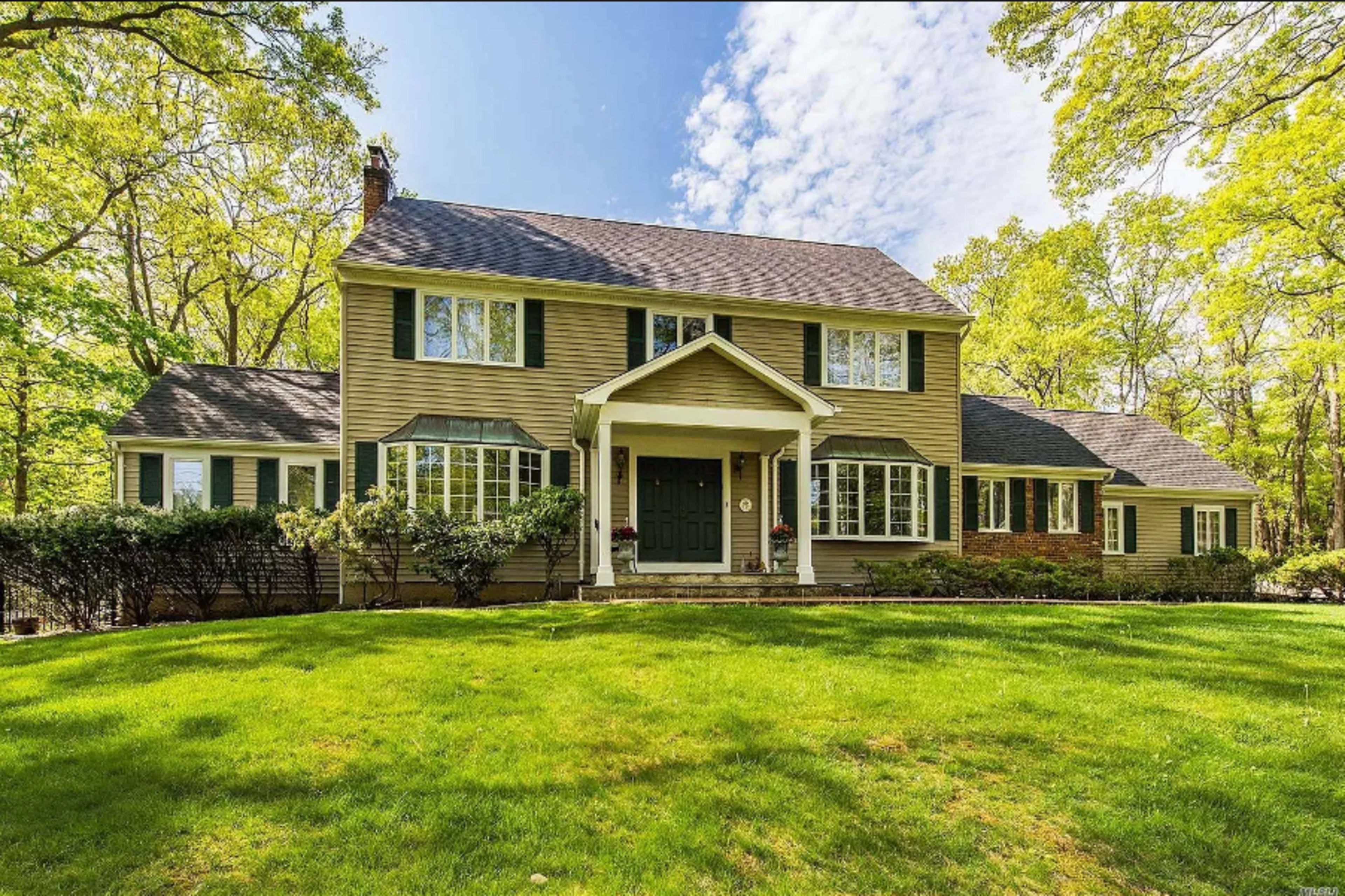 A two-story house with green shutters and a large front lawn is surrounded by trees and features a covered entrance.