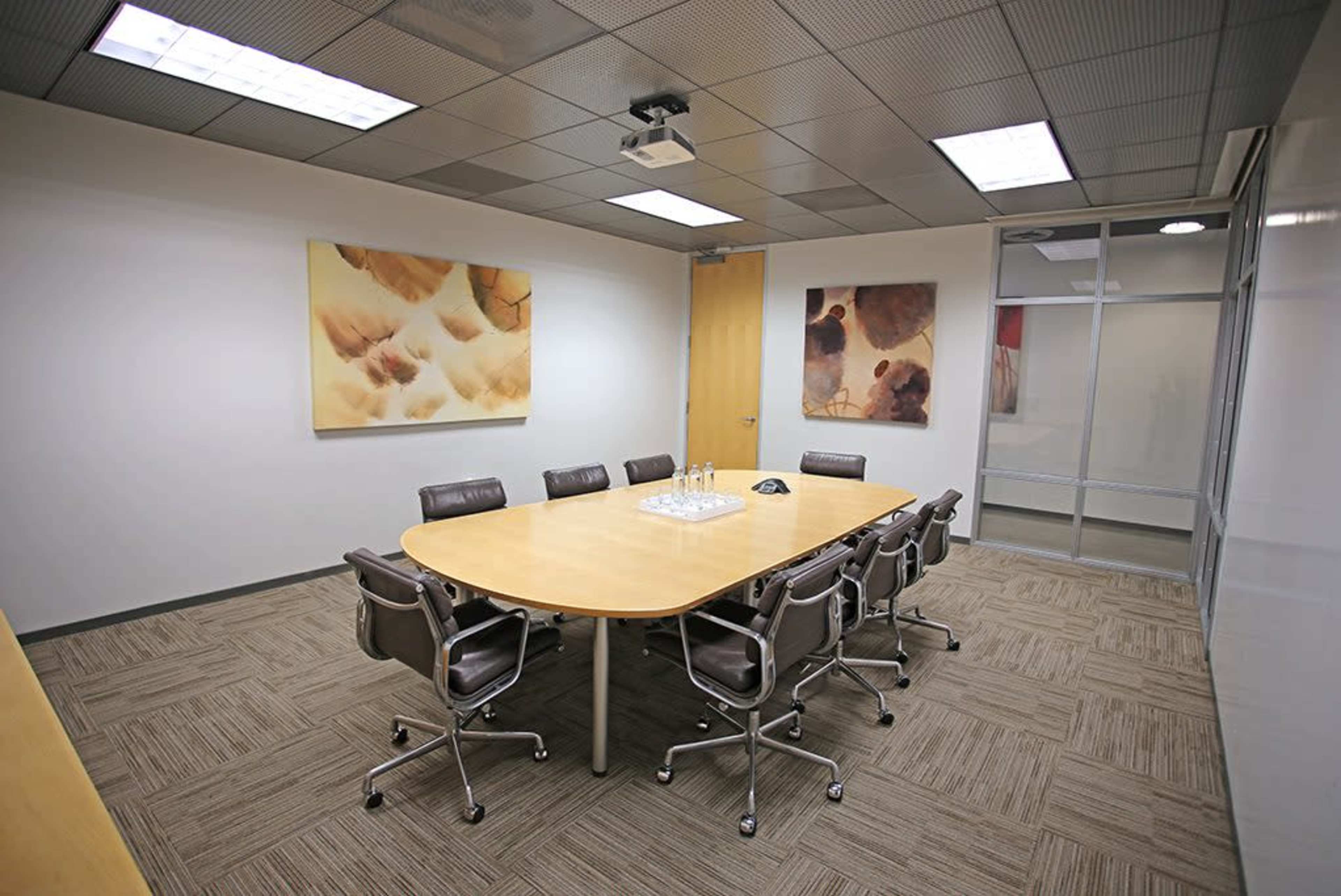 A conference room features a large oval table surrounded by six chairs, with two abstract paintings on the walls and a projector mounted on the ceiling.