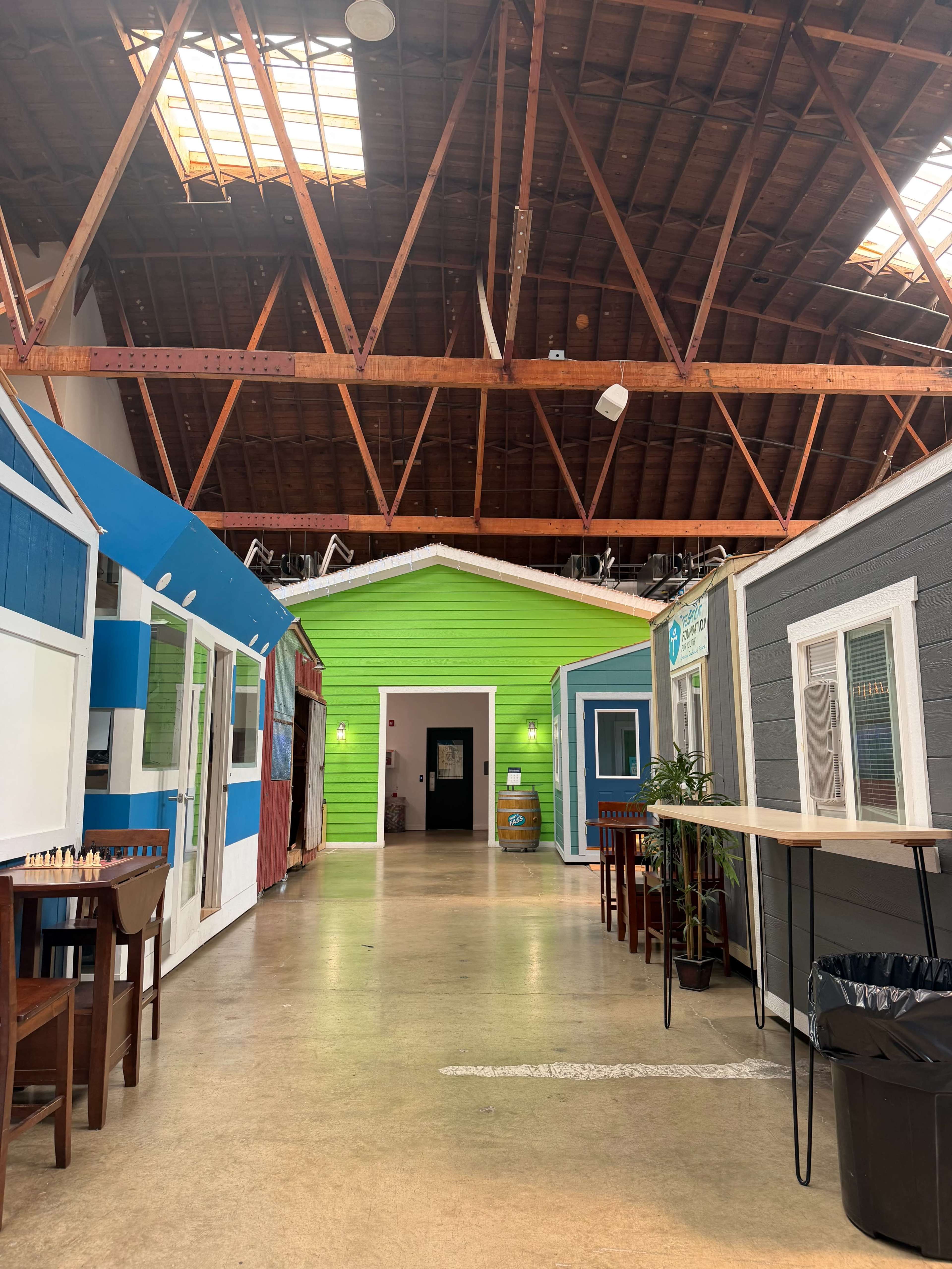 The image shows a hallway featuring colorful house-shaped structures on either side, with a high wooden ceiling and a concrete floor.