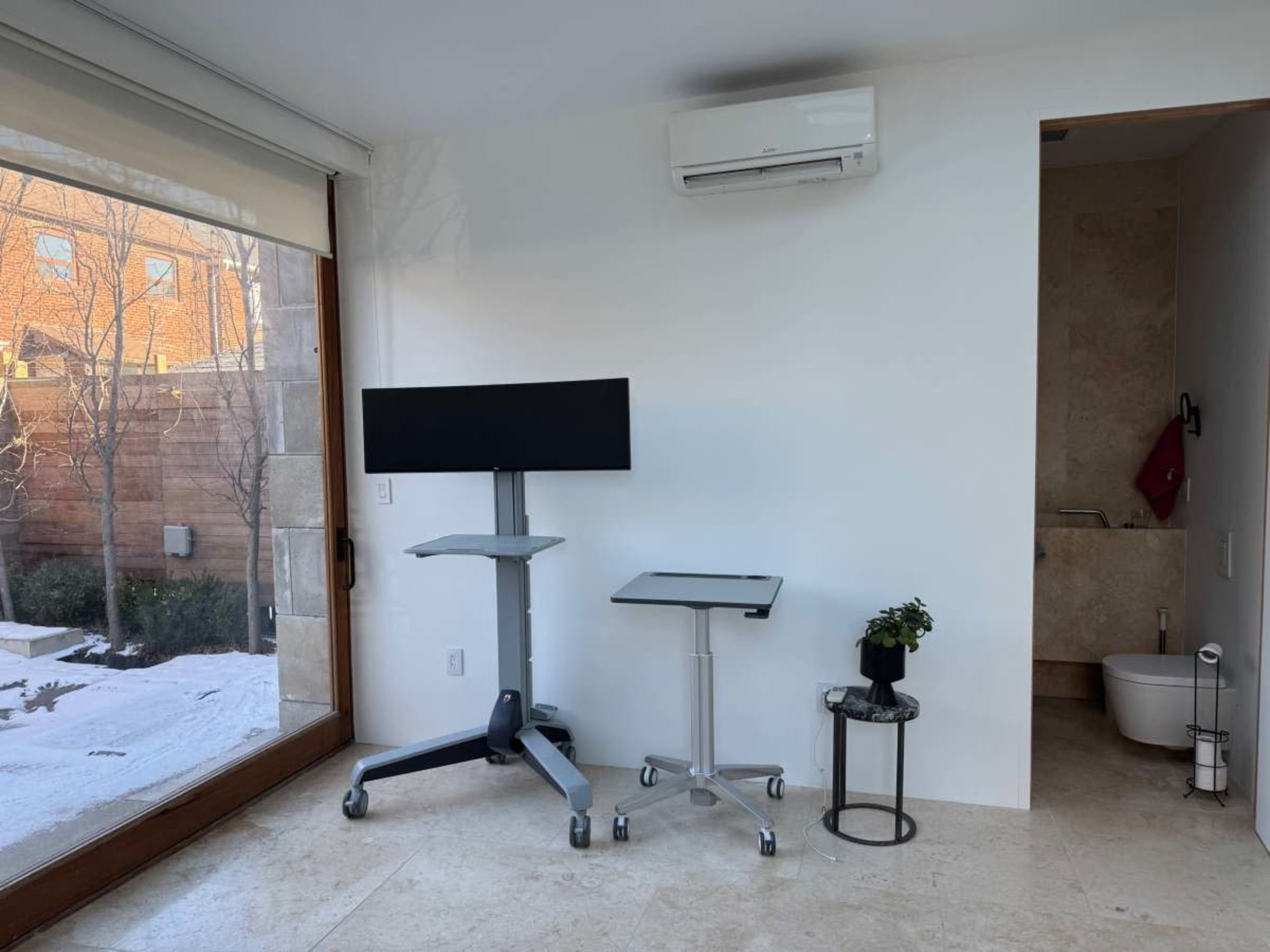 A modern workspace features two movable desks, one with a large monitor, beside a small potted plant and a doorway leading to a bathroom.