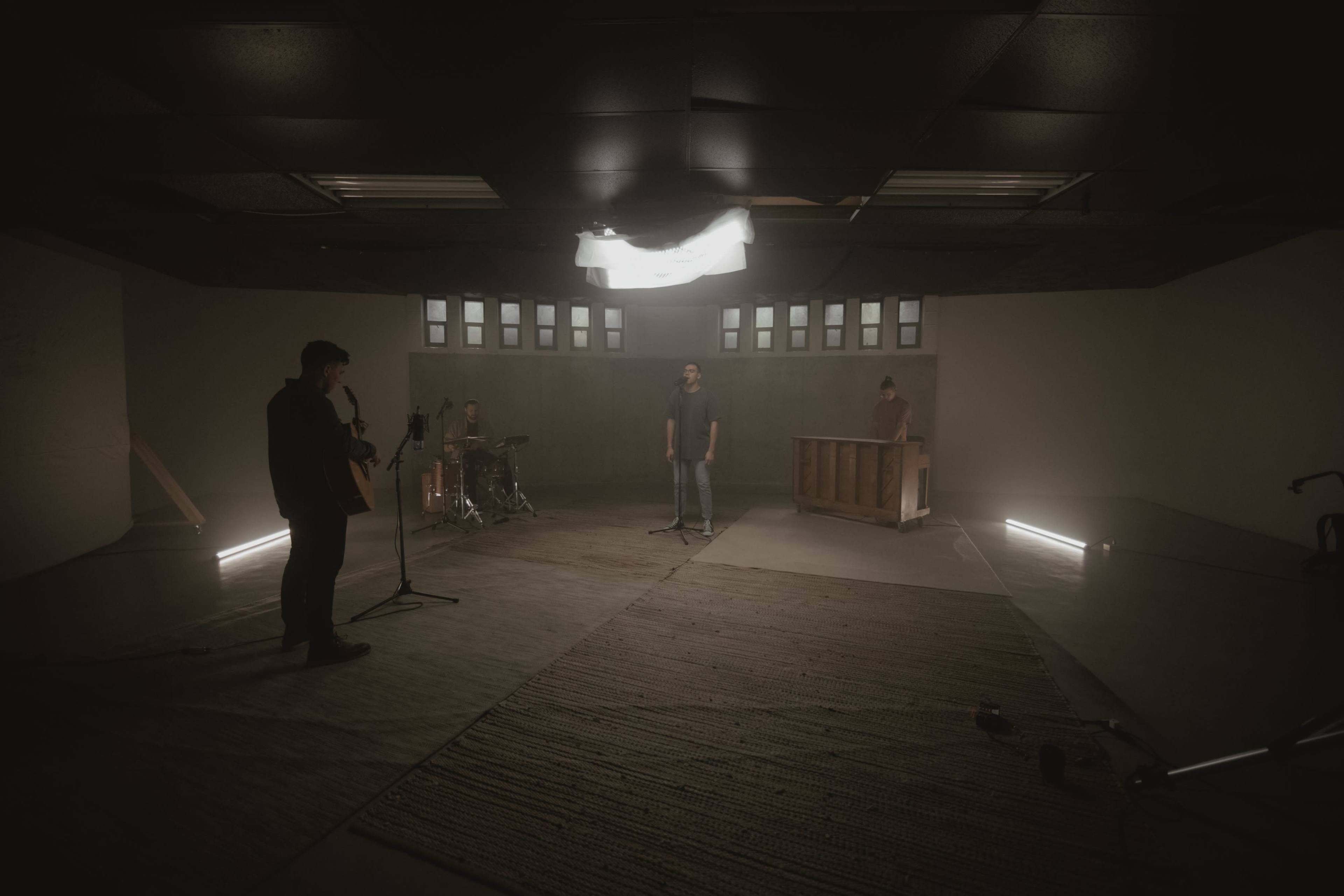 A band performs in a dimly lit rehearsal space featuring instruments like a guitar, drums, and a piano.