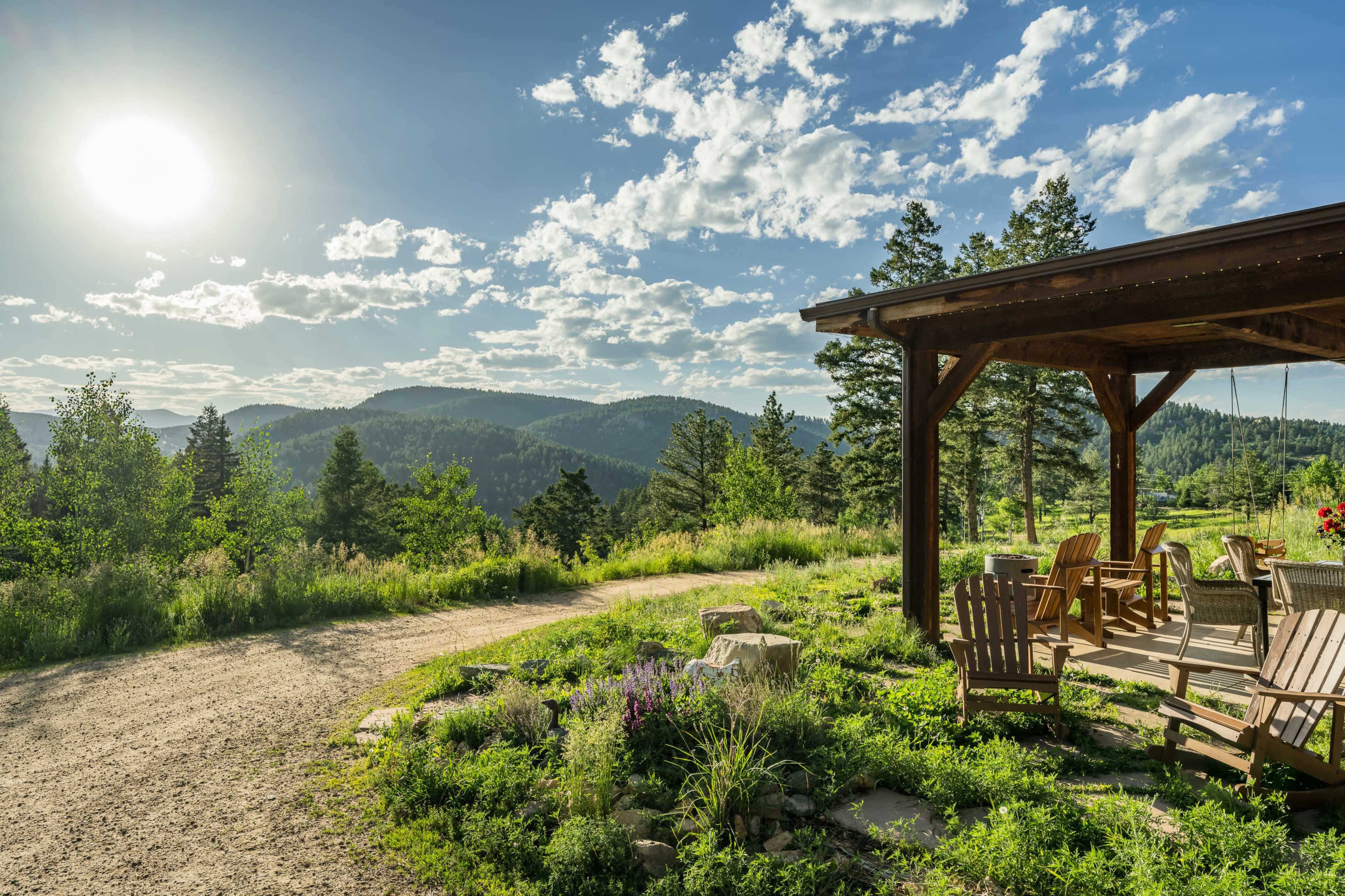 A wooden patio with chairs overlooks a dirt path leading to a mountainous landscape under a blue sky filled with clouds.
