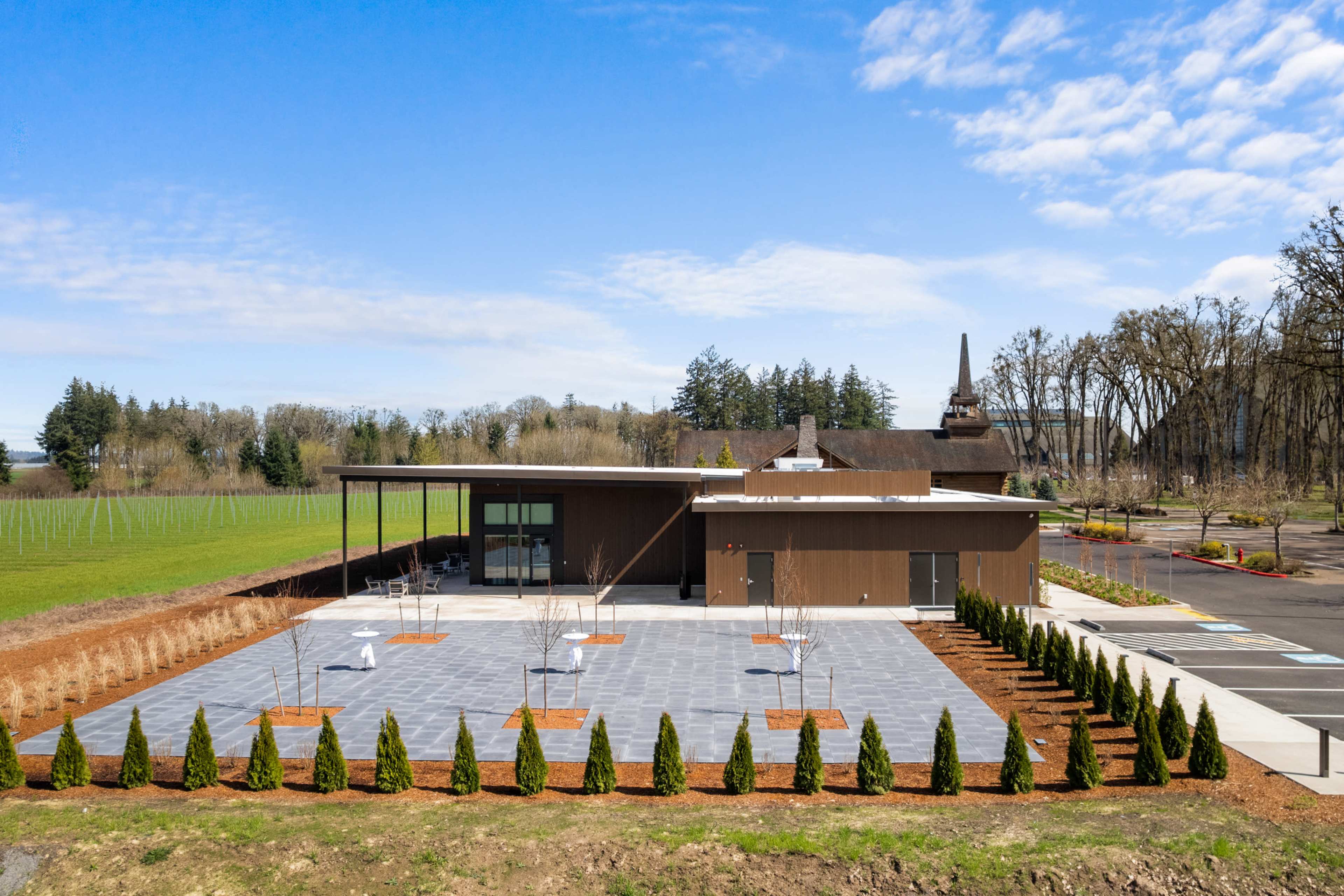 A modern building with a landscaped patio and numerous small trees is set against a rural backdrop of grass and trees under a clear blue sky.