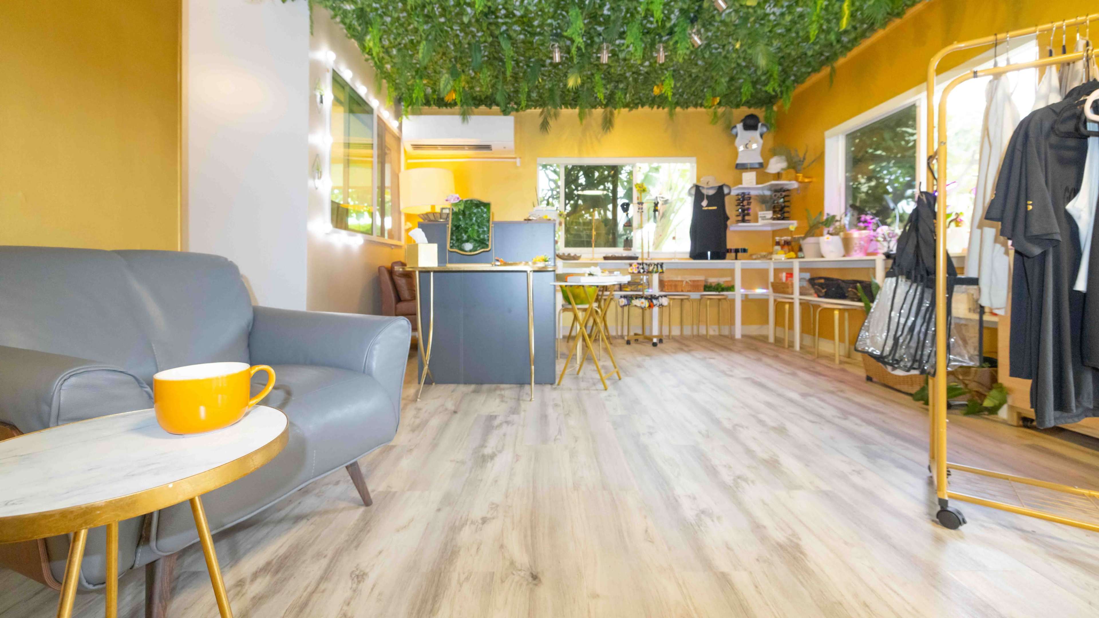 The image shows a cozy interior space with a gray armchair, a small marble-top table, and clothing racks against yellow and white walls, adorned with greenery overhead.