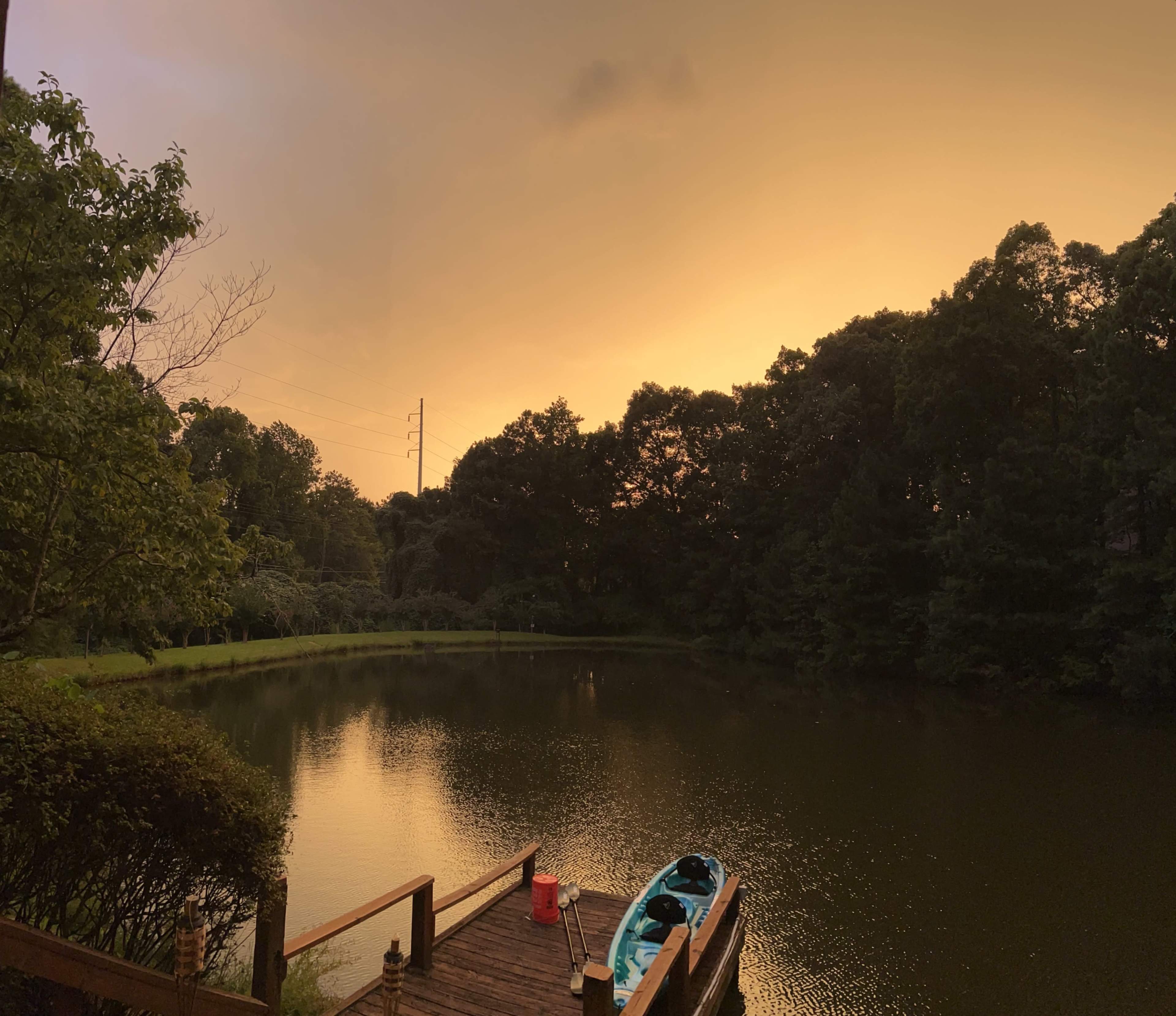 A dock extends into a calm lake surrounded by trees, with a kayak resting on the side and an orange-tinted sky overhead.
