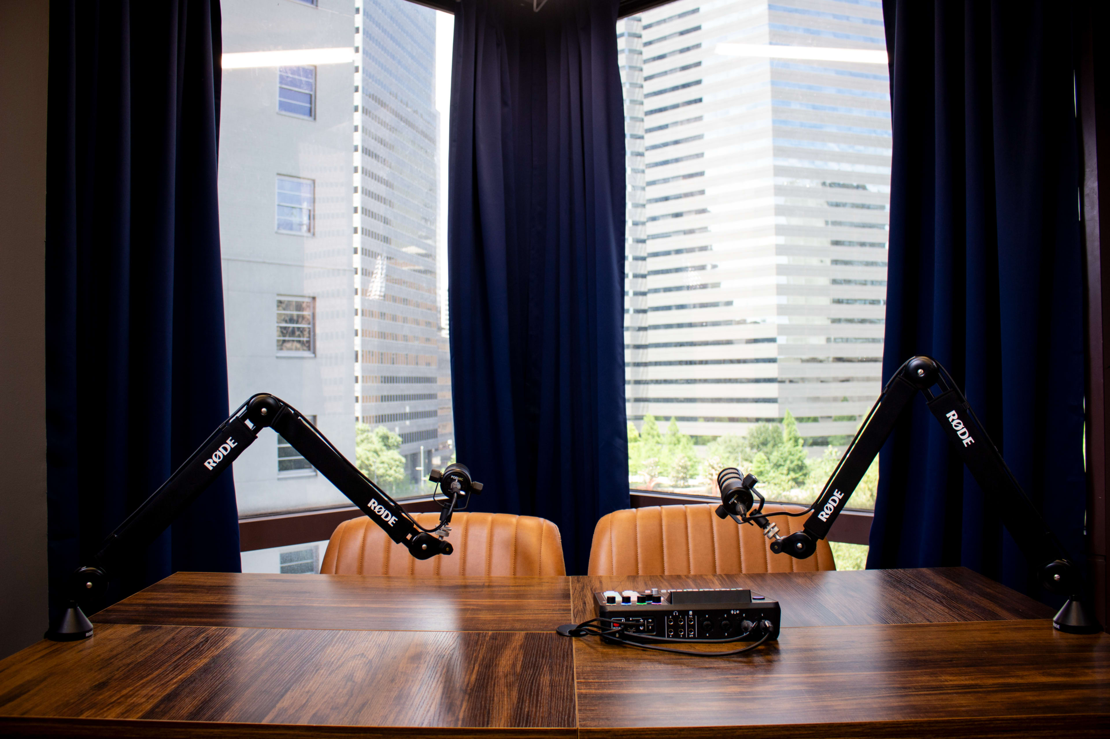 A wooden table with two microphone arms and audio equipment is positioned in front of large windows overlooking city skyscrapers.