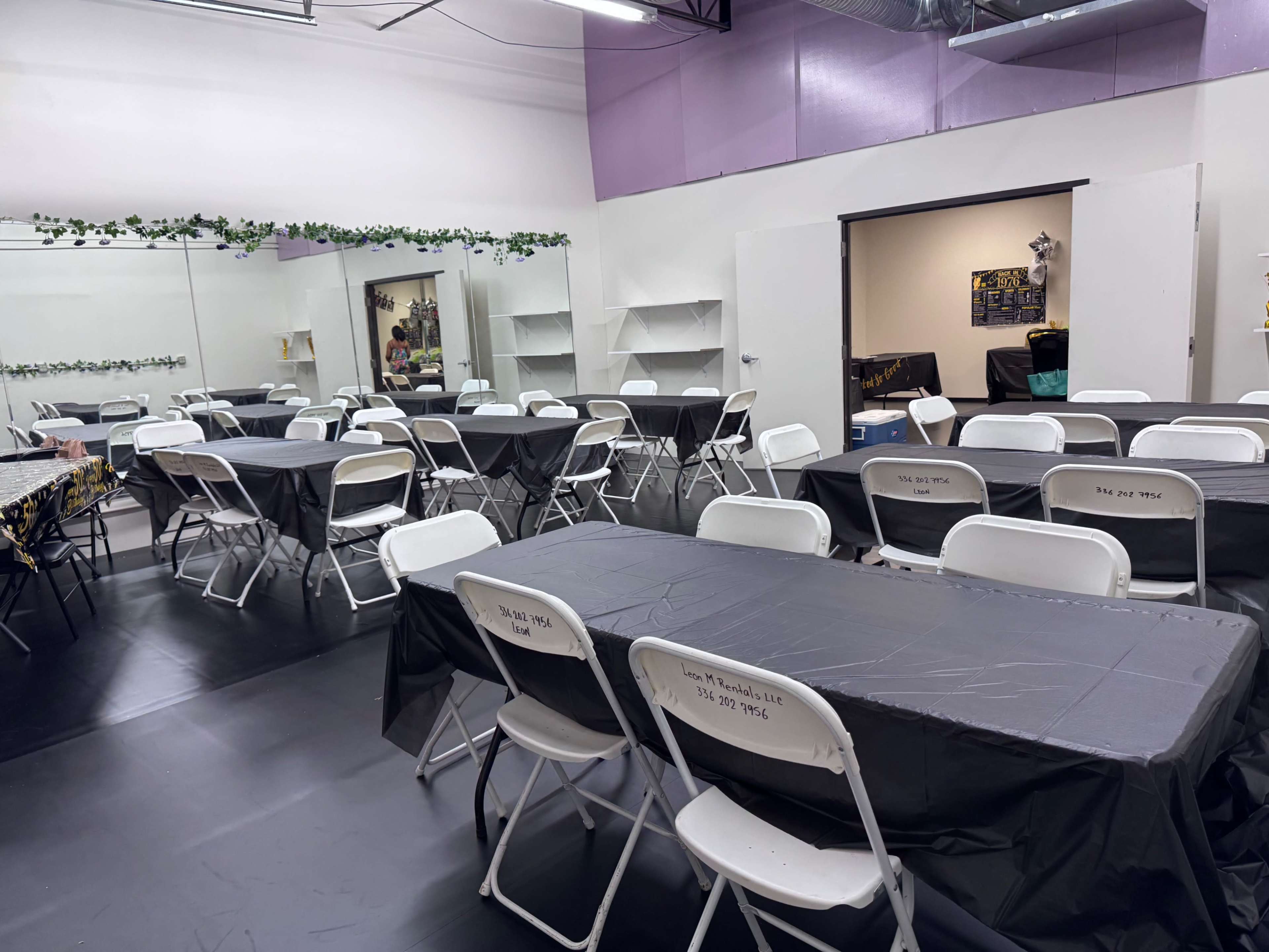 The image shows a spacious room arranged with several tables and chairs covered in black tablecloths, with mirrors on one side and shelving in the background.