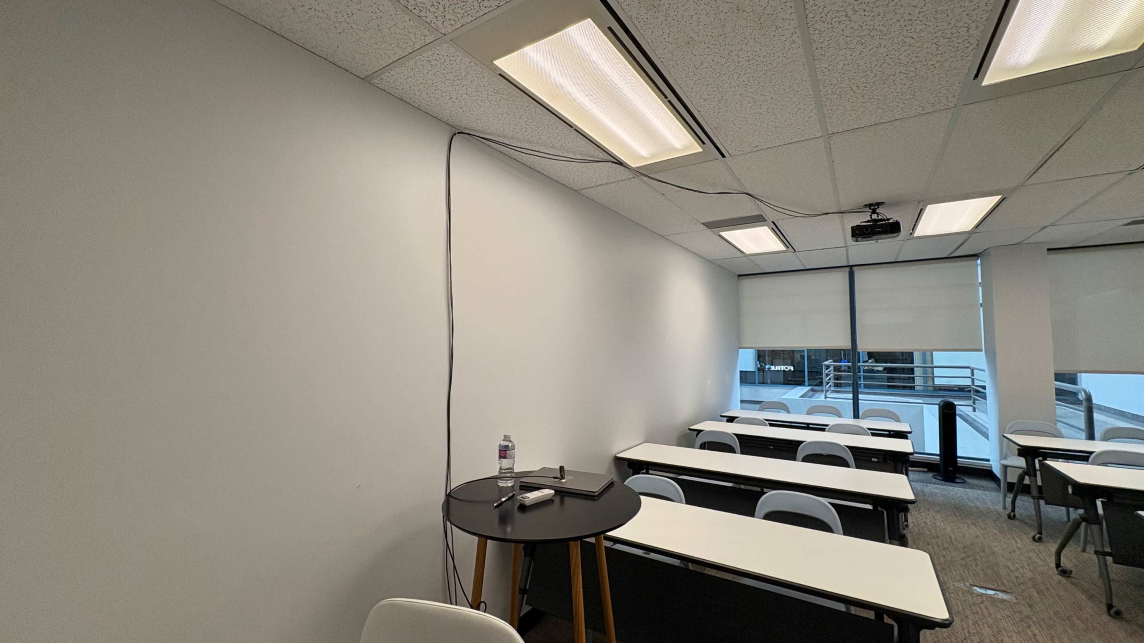 A classroom is shown with white walls, rows of tables and chairs, and a projector mounted on the ceiling.