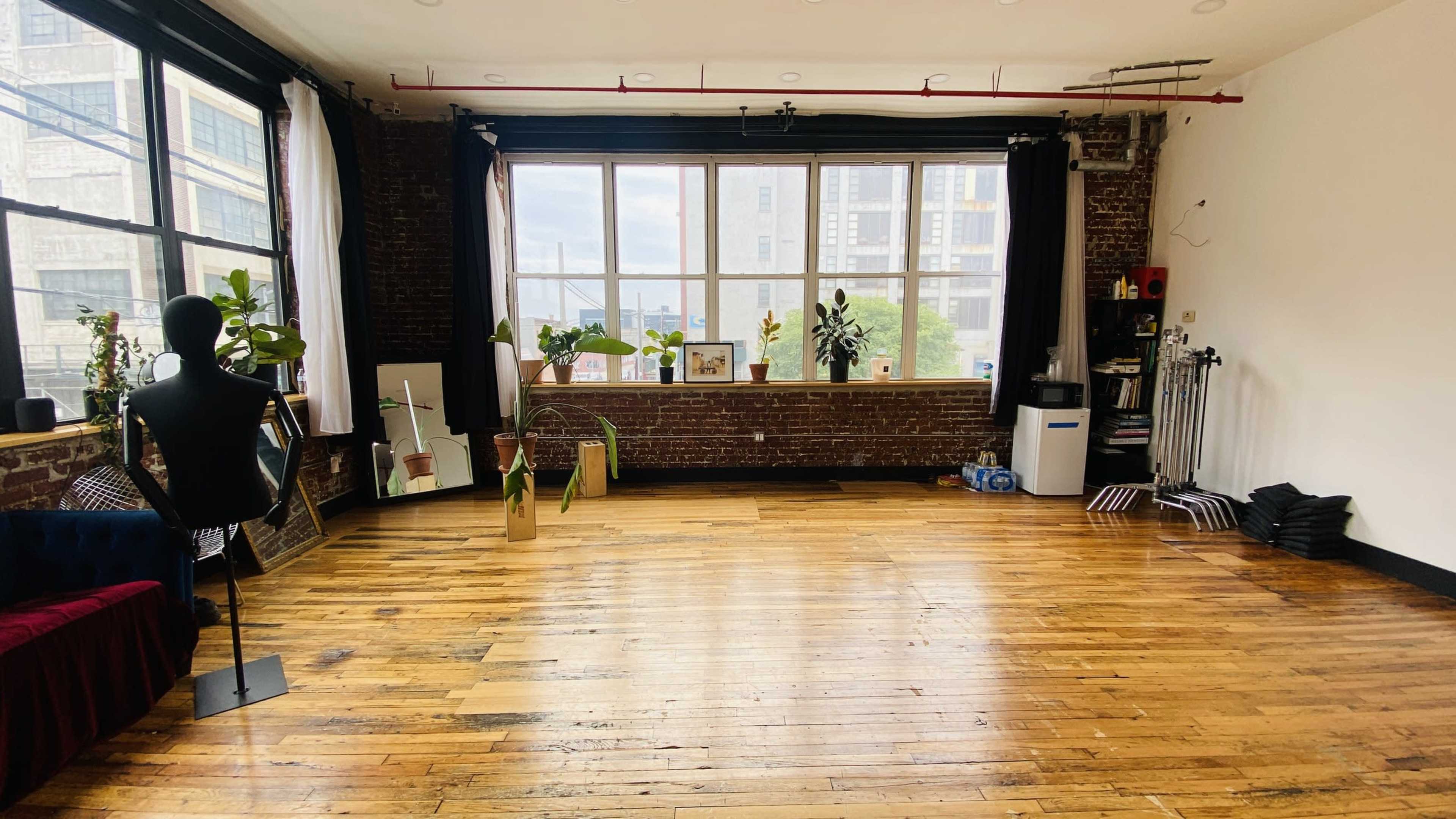 The image shows a spacious room with wooden flooring, large windows, and various potted plants arranged around the space.