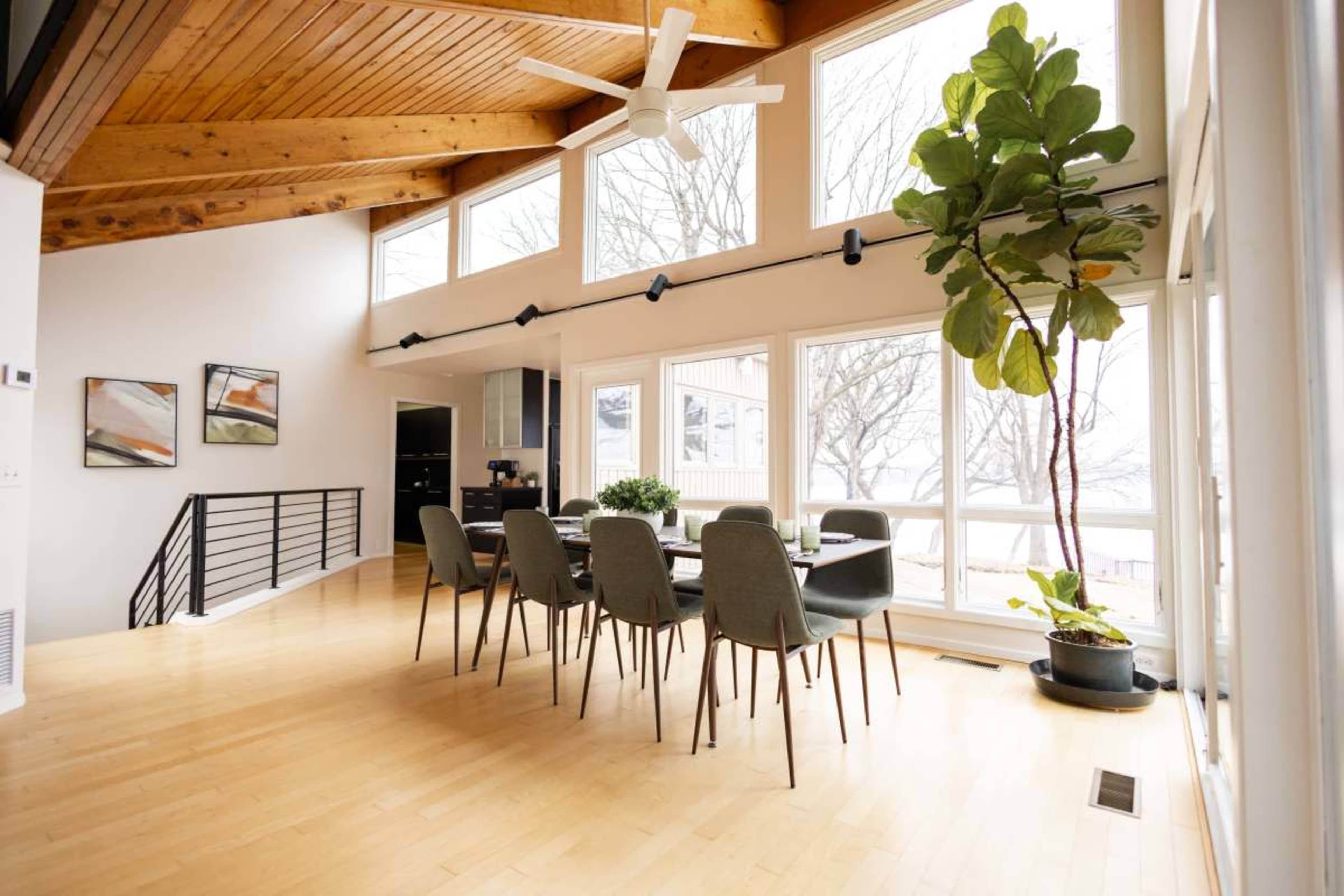 A spacious dining area features a wooden ceiling, large windows, and a table set for six, accompanied by a potted plant in the corner.