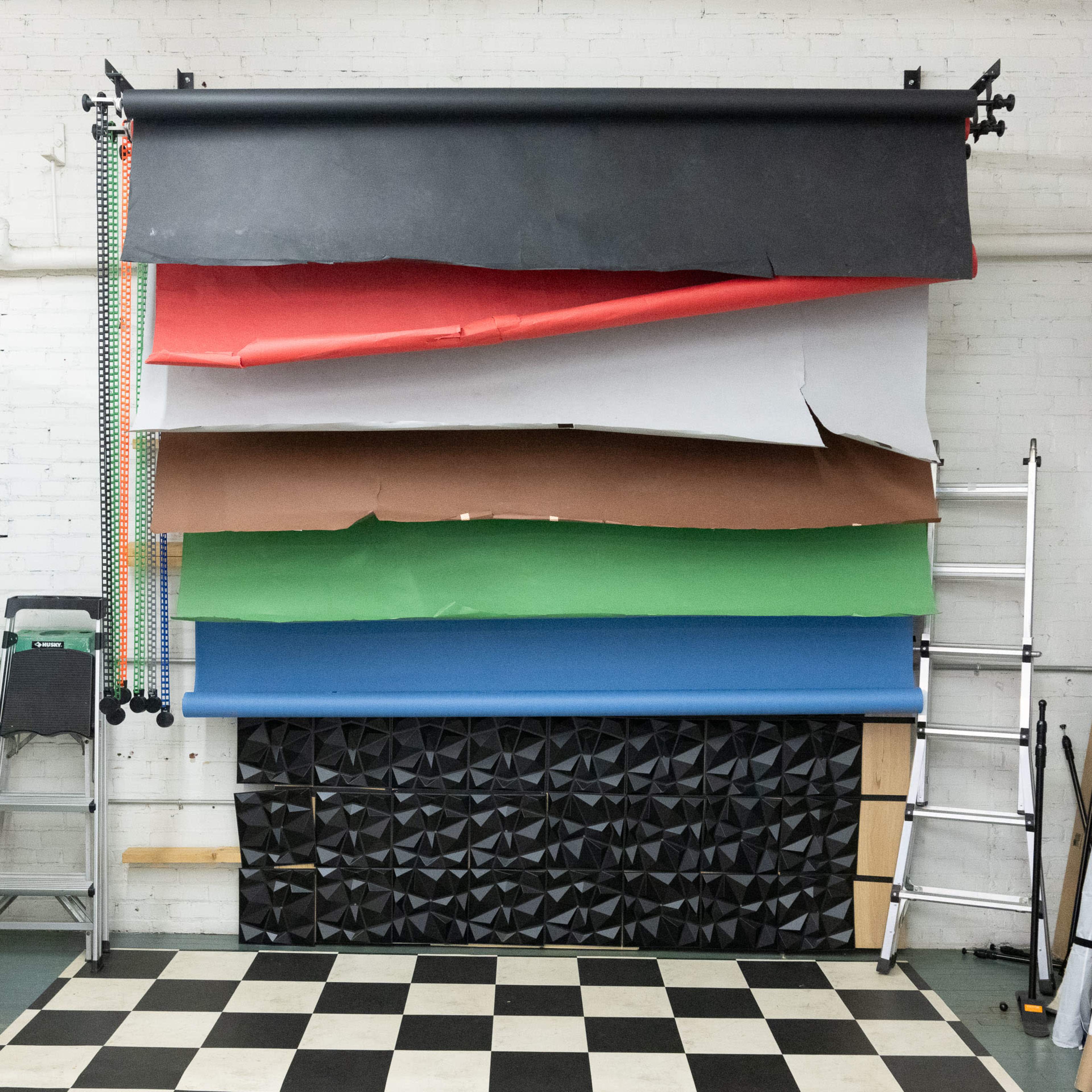 A studio backdrop setup displays several rolls of colored paper stacked vertically against a wall, with a ladder leaning nearby on a checkered floor.
