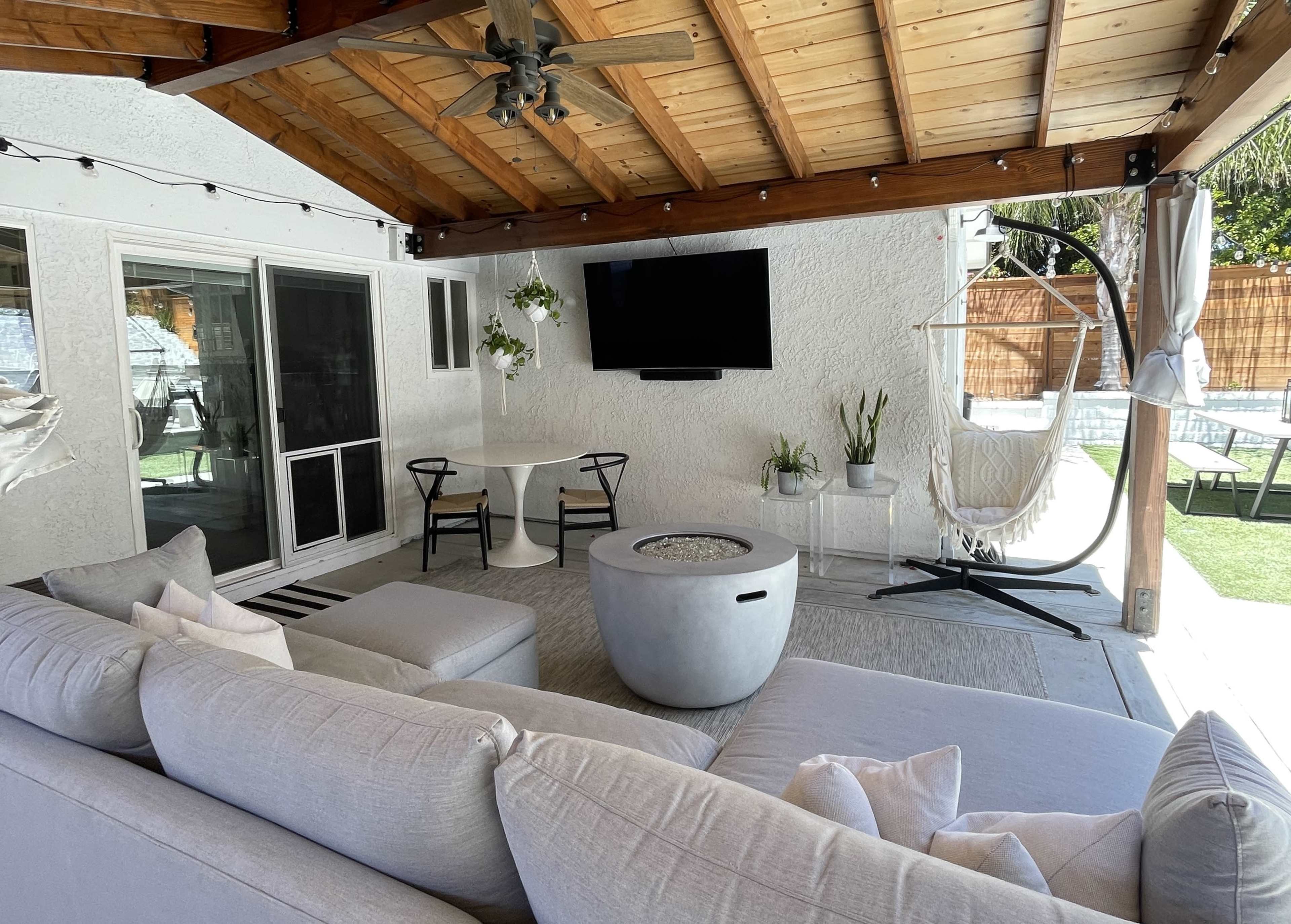 A covered outdoor living area featuring a gray sectional sofa, a round table, a television mounted on the wall, and a hanging chair.