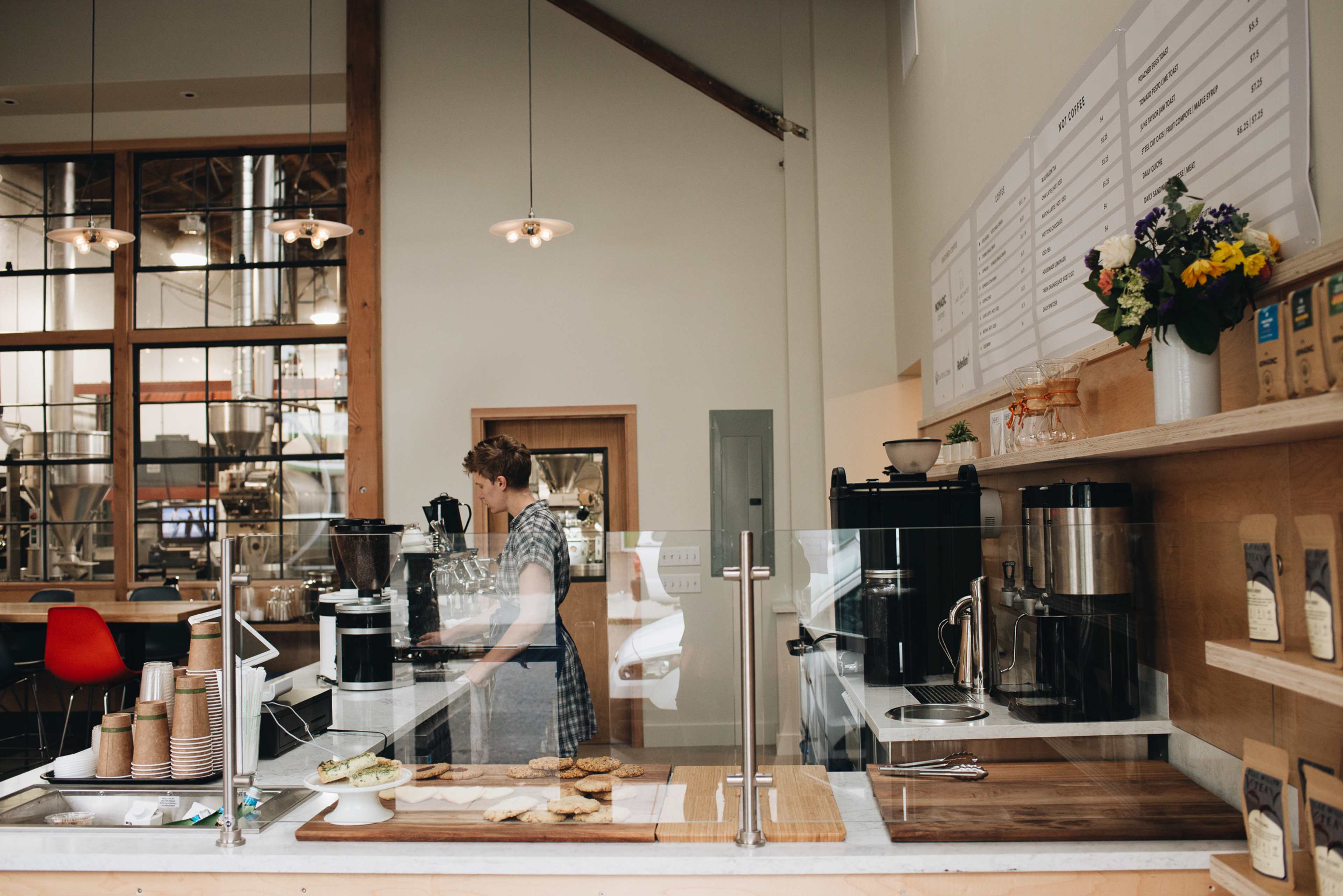 A barista prepares coffee behind a glass counter in a modern café featuring wooden accents and baked goods on display.