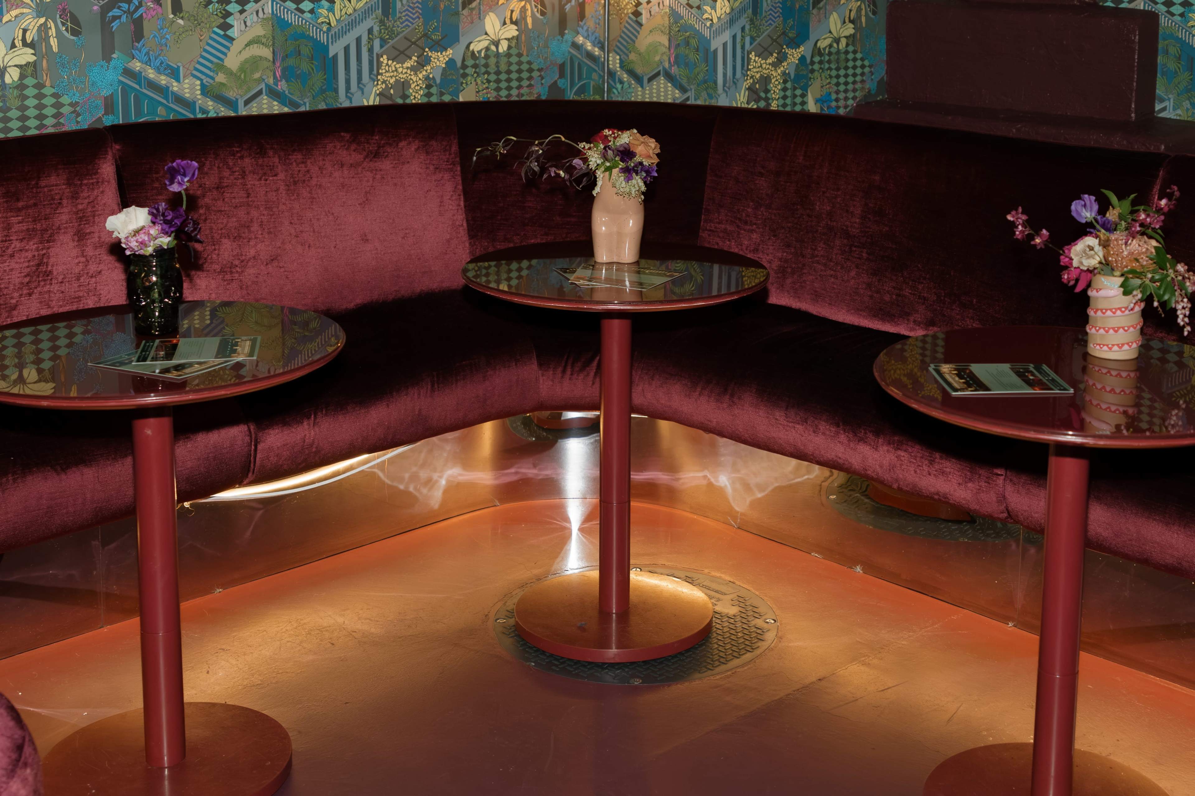 The image shows a cozy seating area with maroon velvet couches, two round tables, and small vases containing flowers.