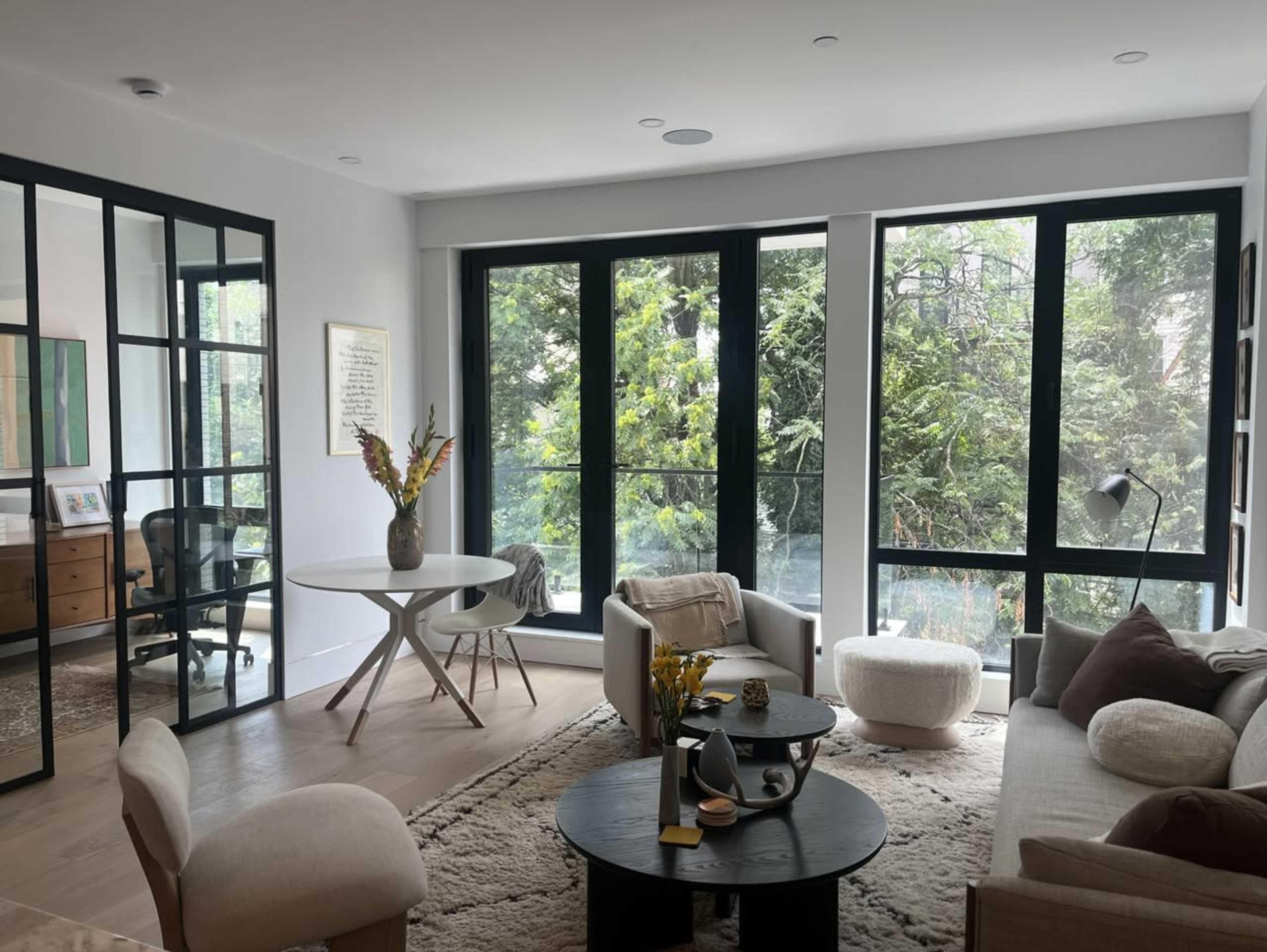 The image features a modern living room with large windows, a circular dining table, and a combination of plush seating arranged around a coffee table on a textured rug.