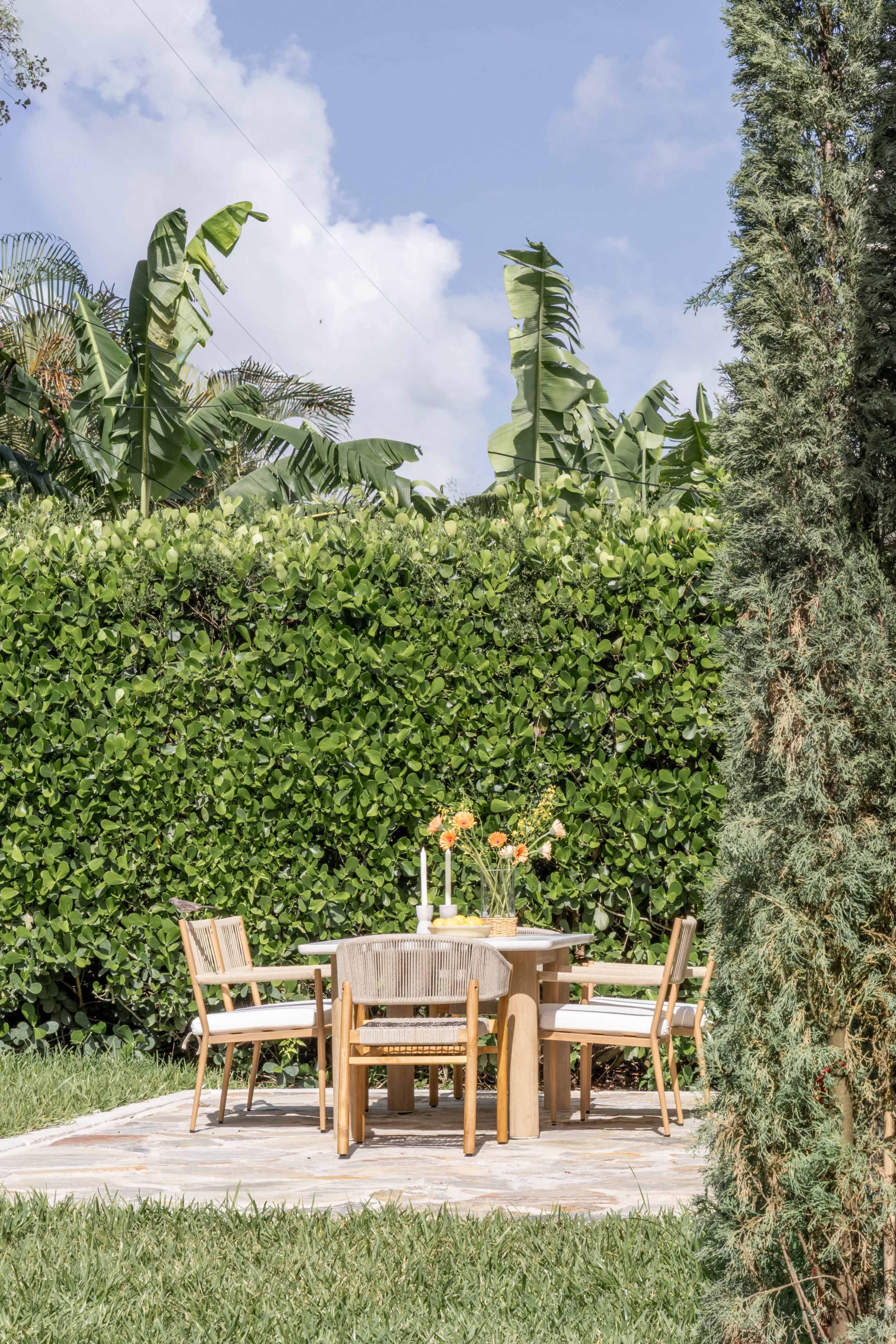 A wooden dining table with chairs is arranged on a stone patio surrounded by lush green foliage and banana plants.