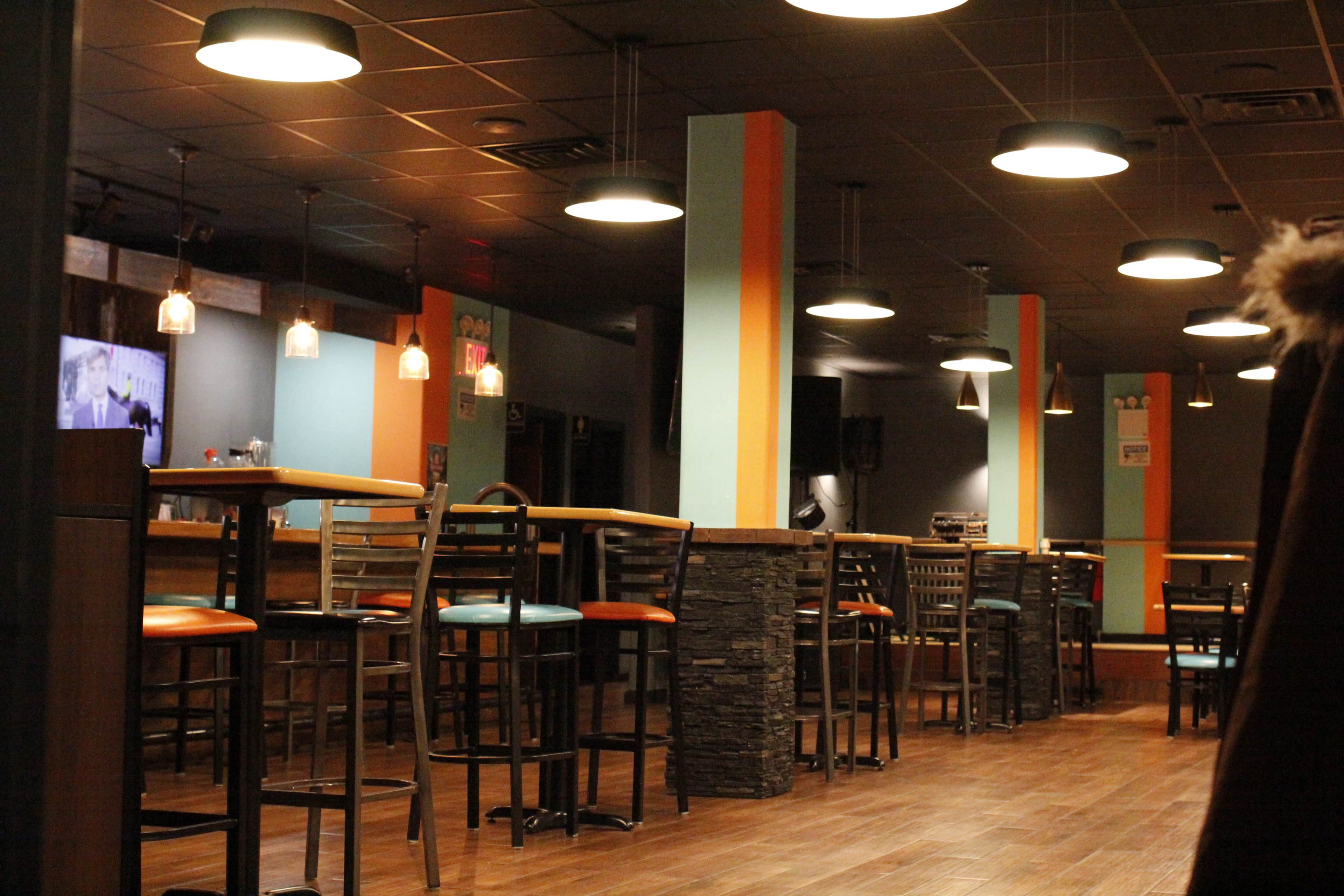 The interior of a bar with high stools and tables, illuminated by pendant lights.