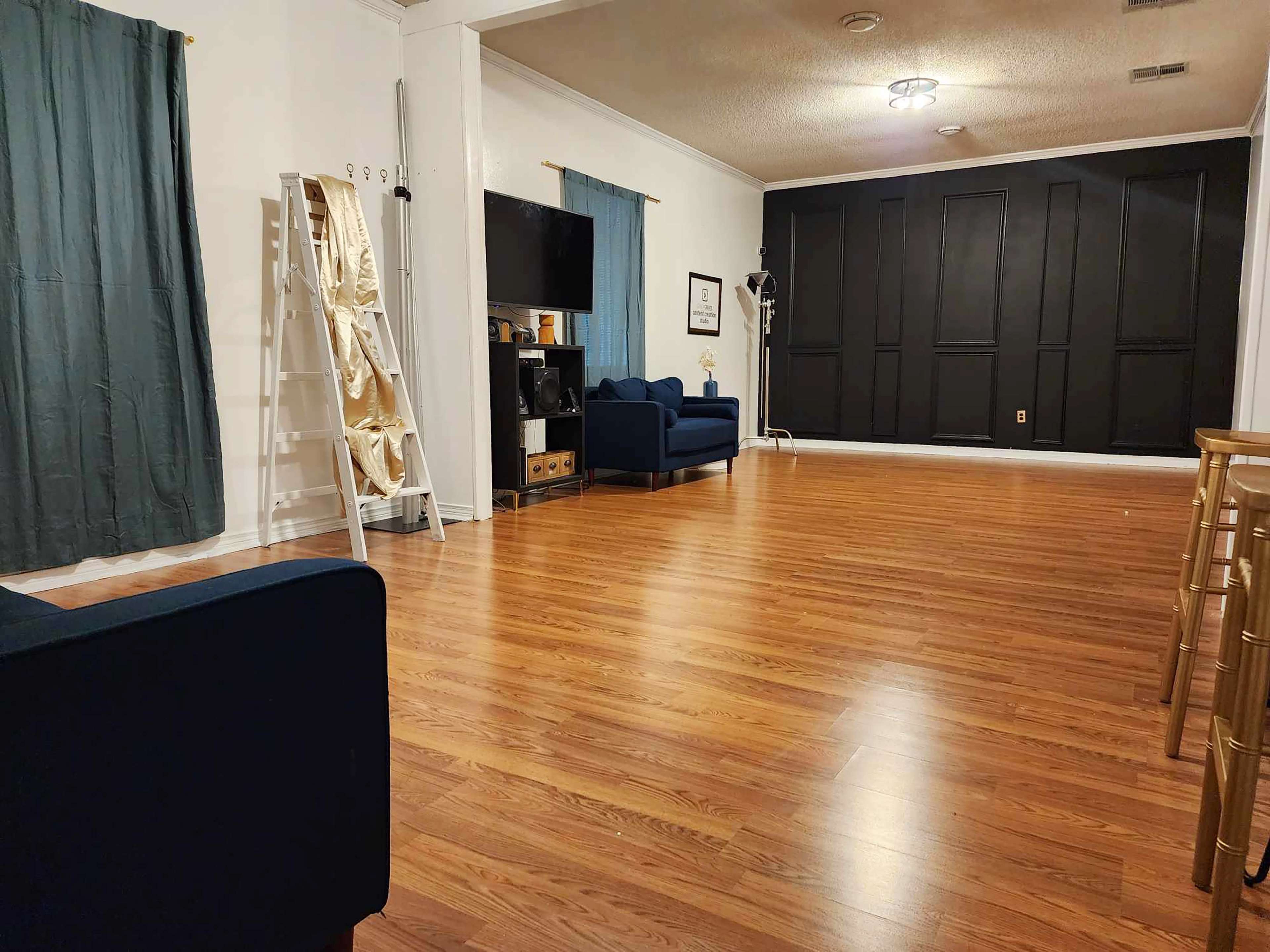 The room features wooden flooring, two blue couches, a black accent wall, and tall windows covered with curtains.