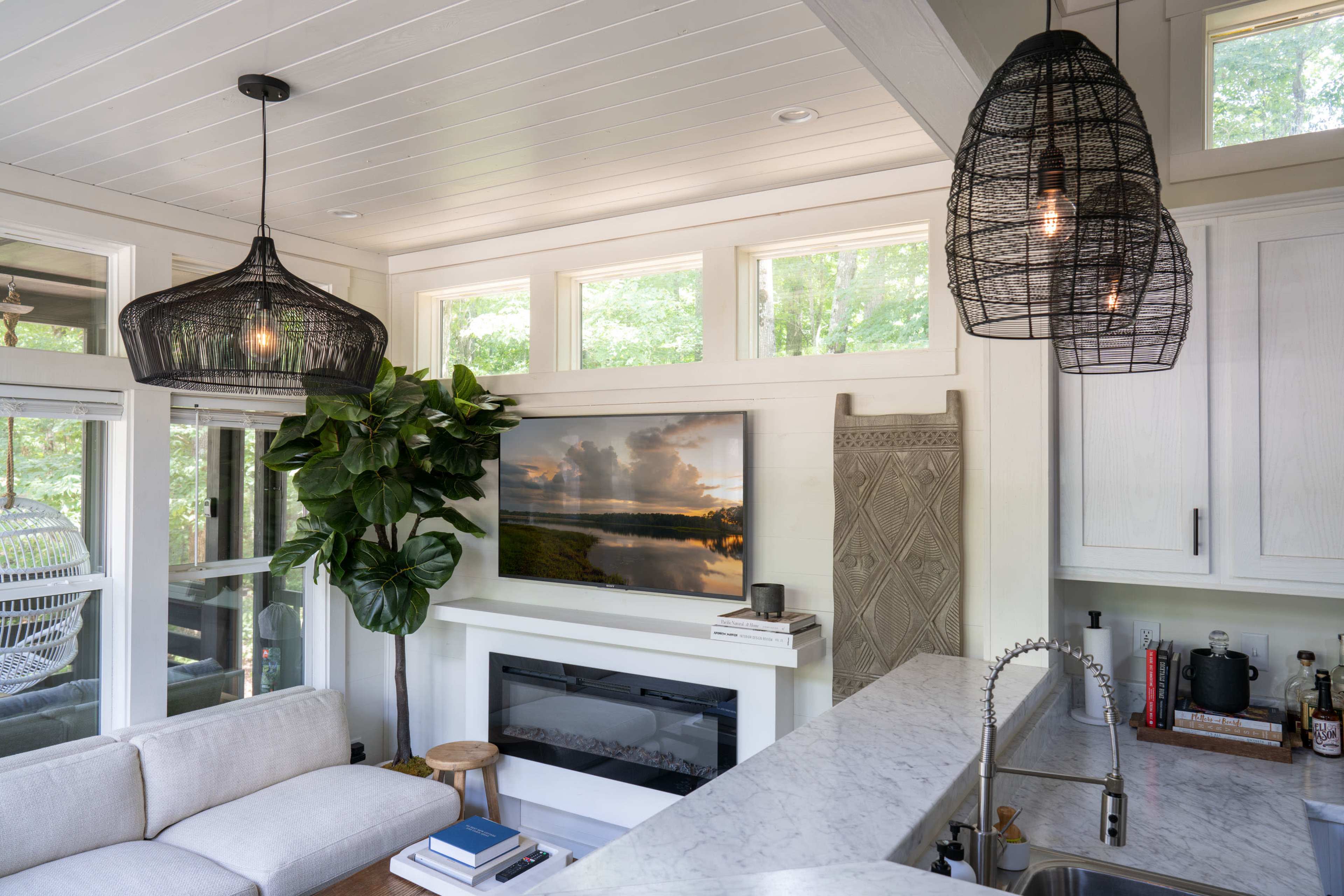 A cozy living room features a large television above a marble fireplace, surrounded by large windows and two pendant lights hanging from the ceiling.