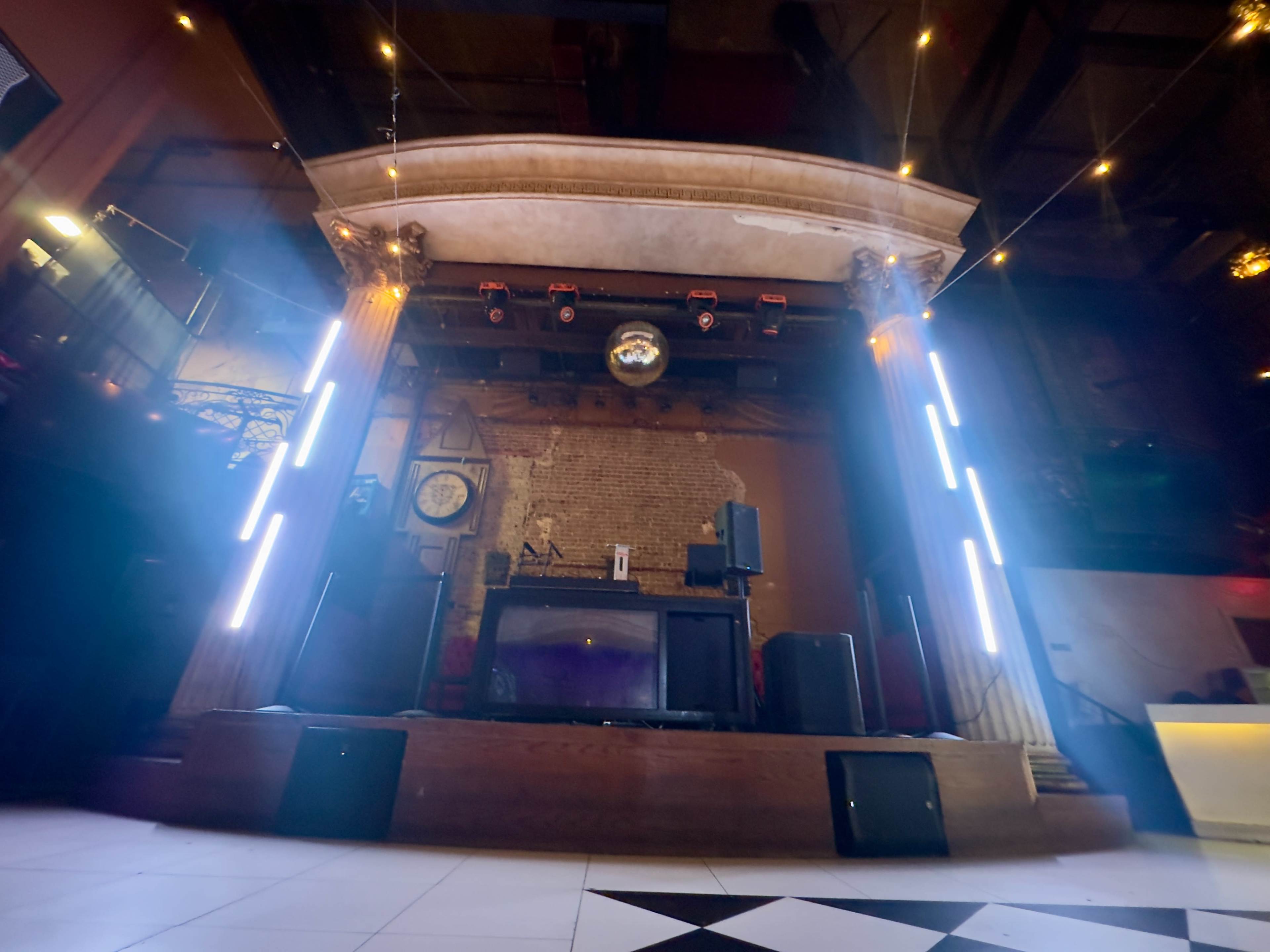 The image shows a stage in a music venue with a disco ball, speakers, and illuminated columns in a dimly lit room.