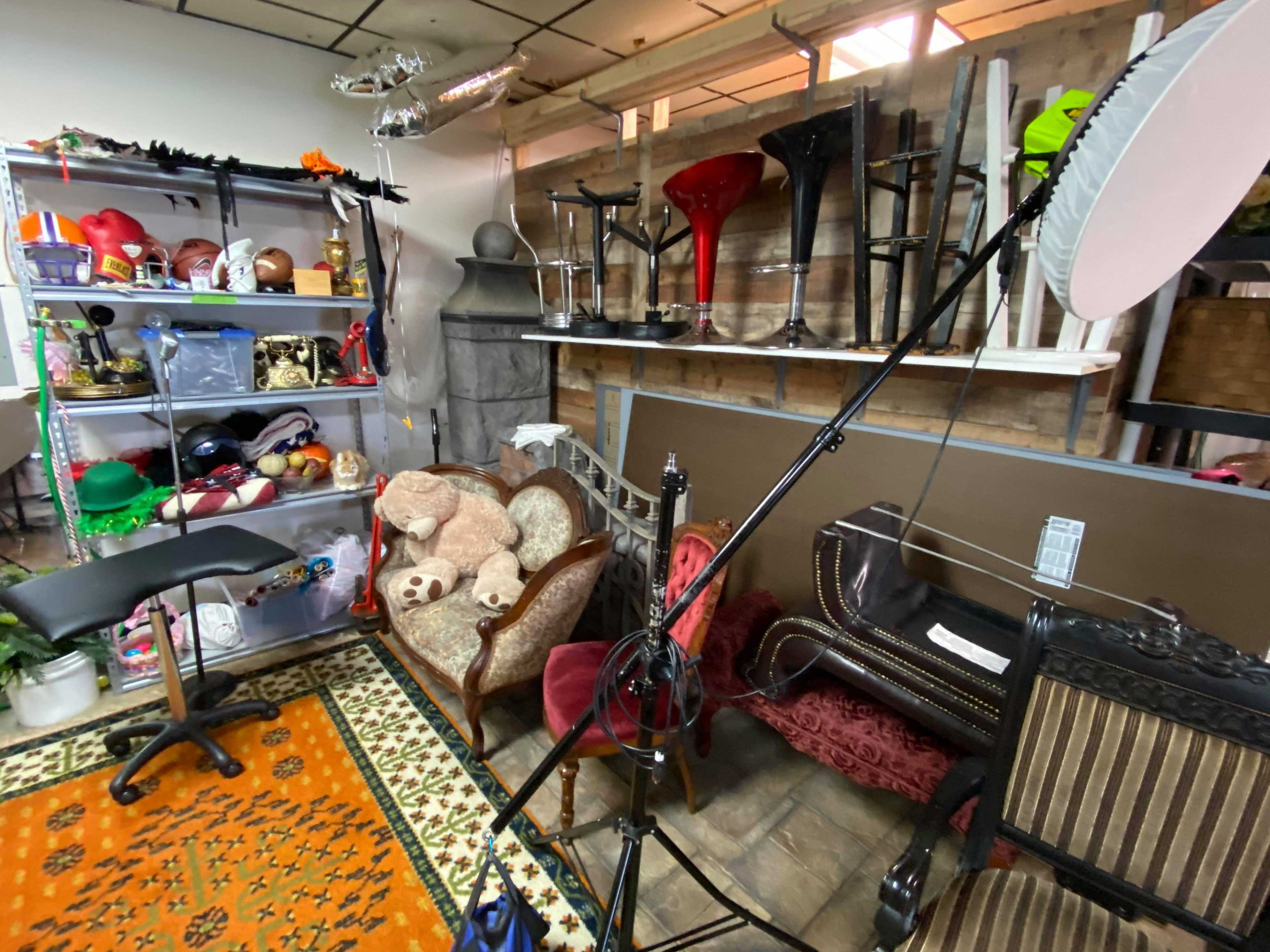 The image shows a cluttered storage area with various props, furniture, and equipment, including chairs, a table, and lighting equipment.