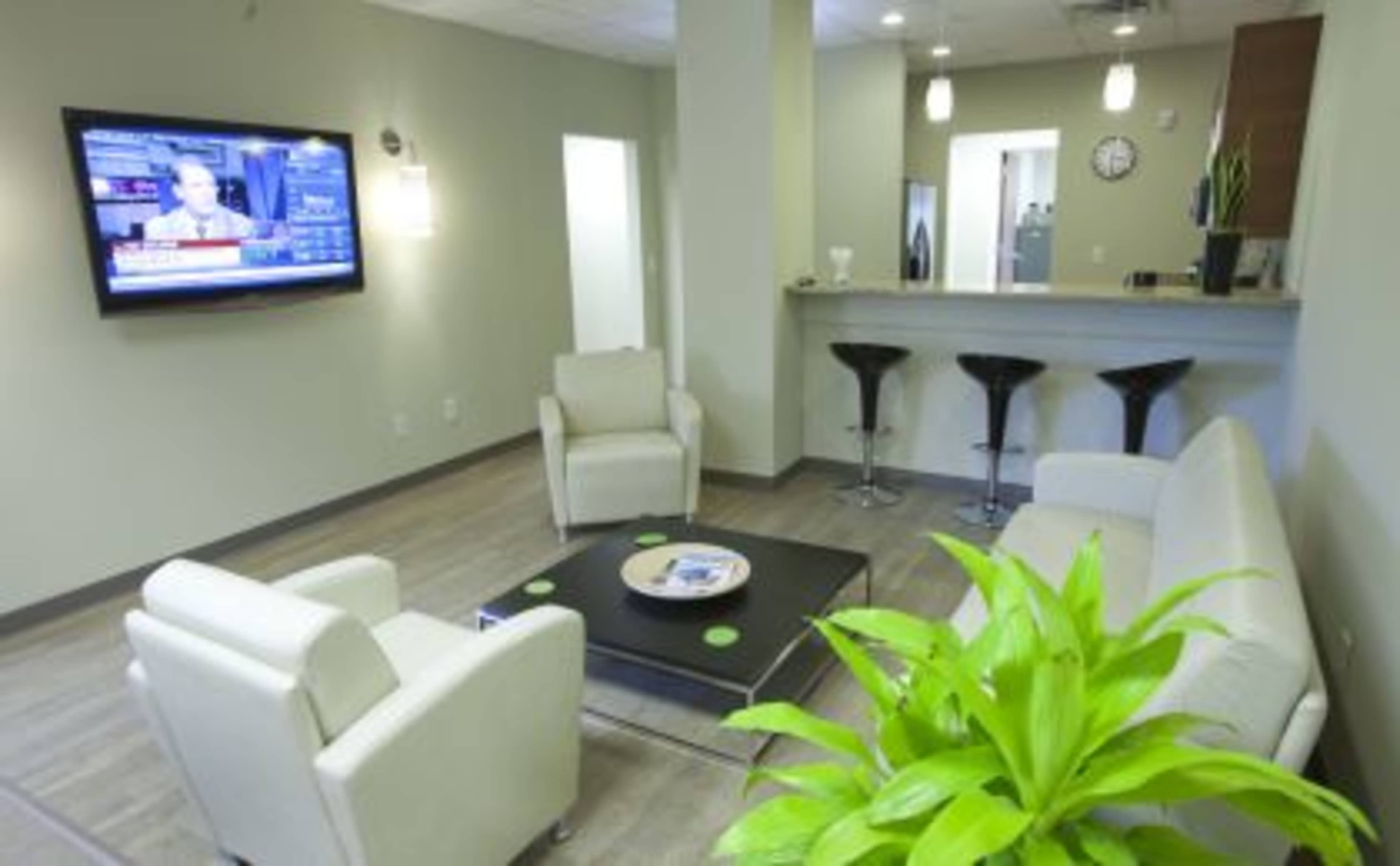 A modern waiting area features two white chairs and a black coffee table next to a television displaying news, with a bar area in the background and a large green plant in the foreground.
