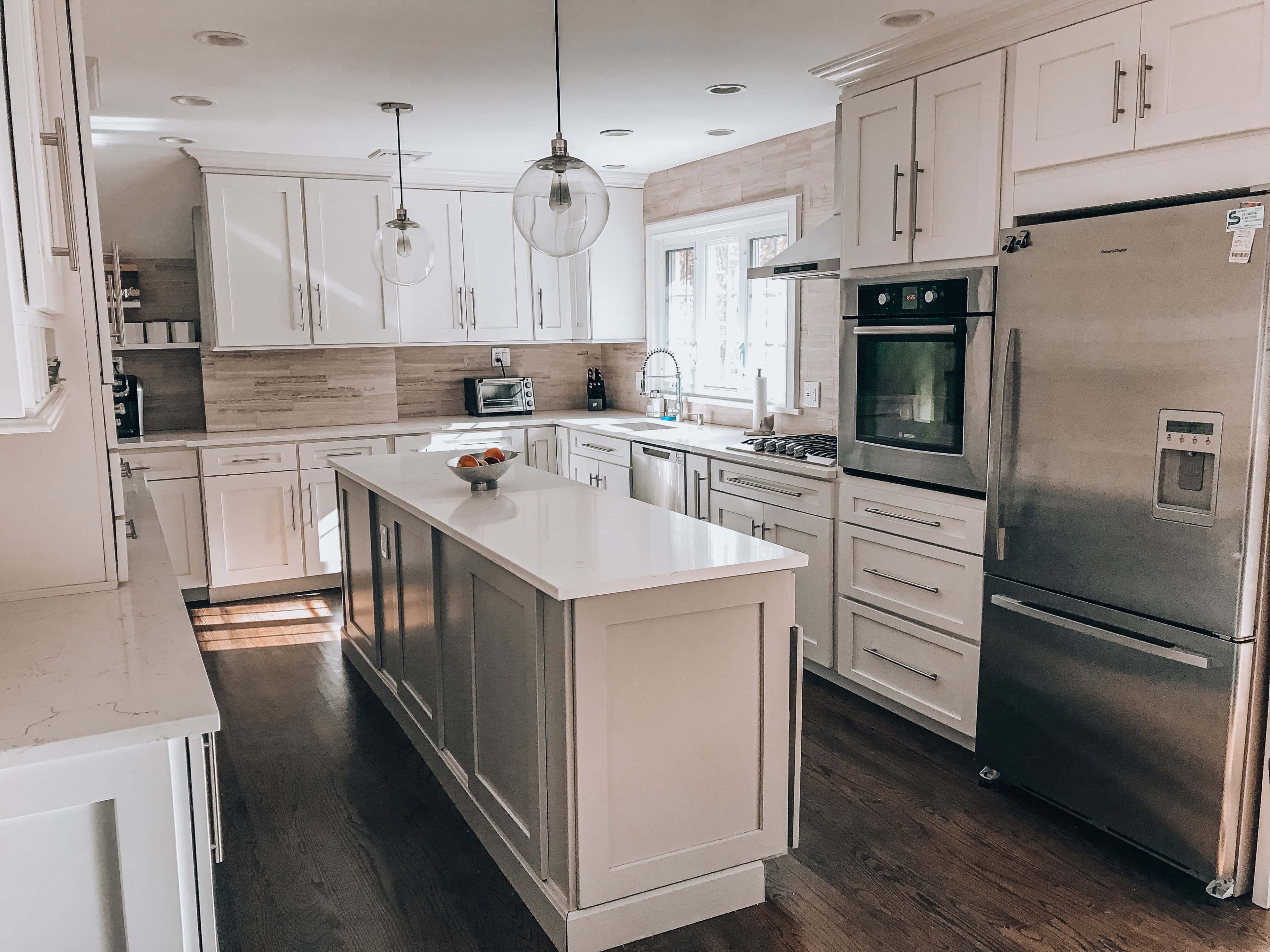 A modern kitchen with white cabinets, a central island, stainless steel appliances, and a large window providing natural light.