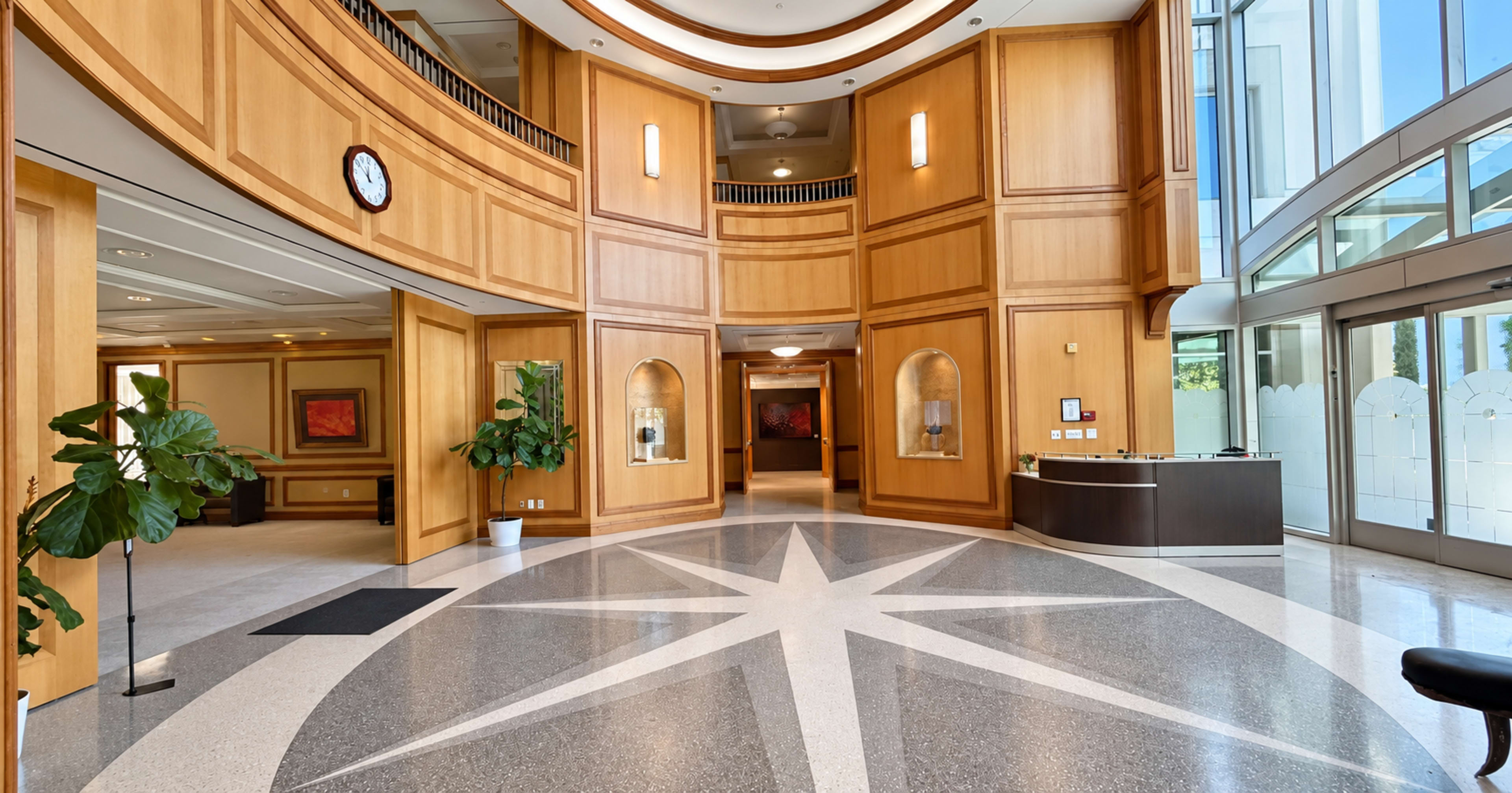 The image shows a spacious lobby with wooden paneling, a star-patterned floor, and large windows allowing natural light to enter.
