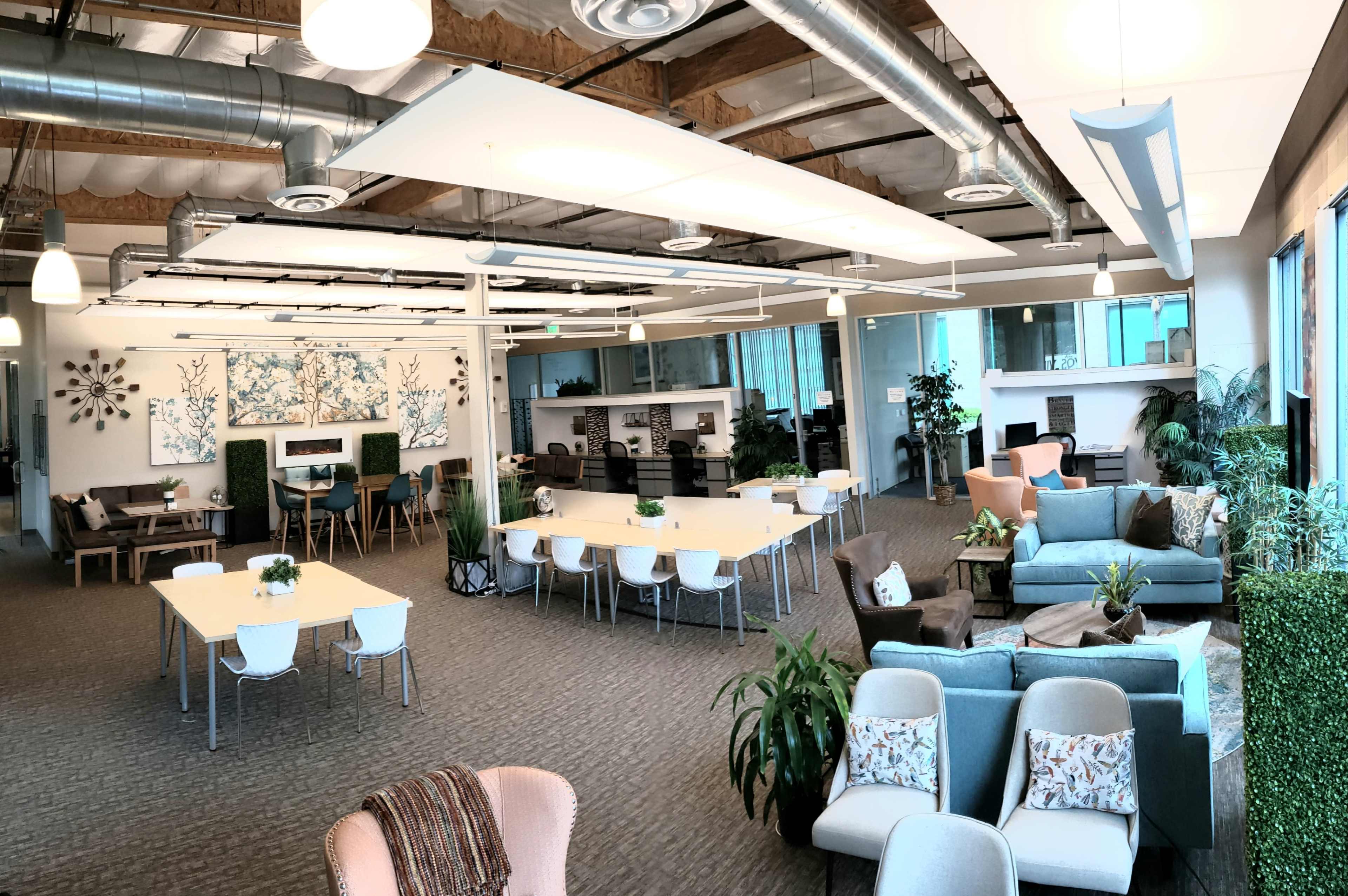 An open office space with various seating areas, tables, and plants, featuring both collaborative and individual workspaces.