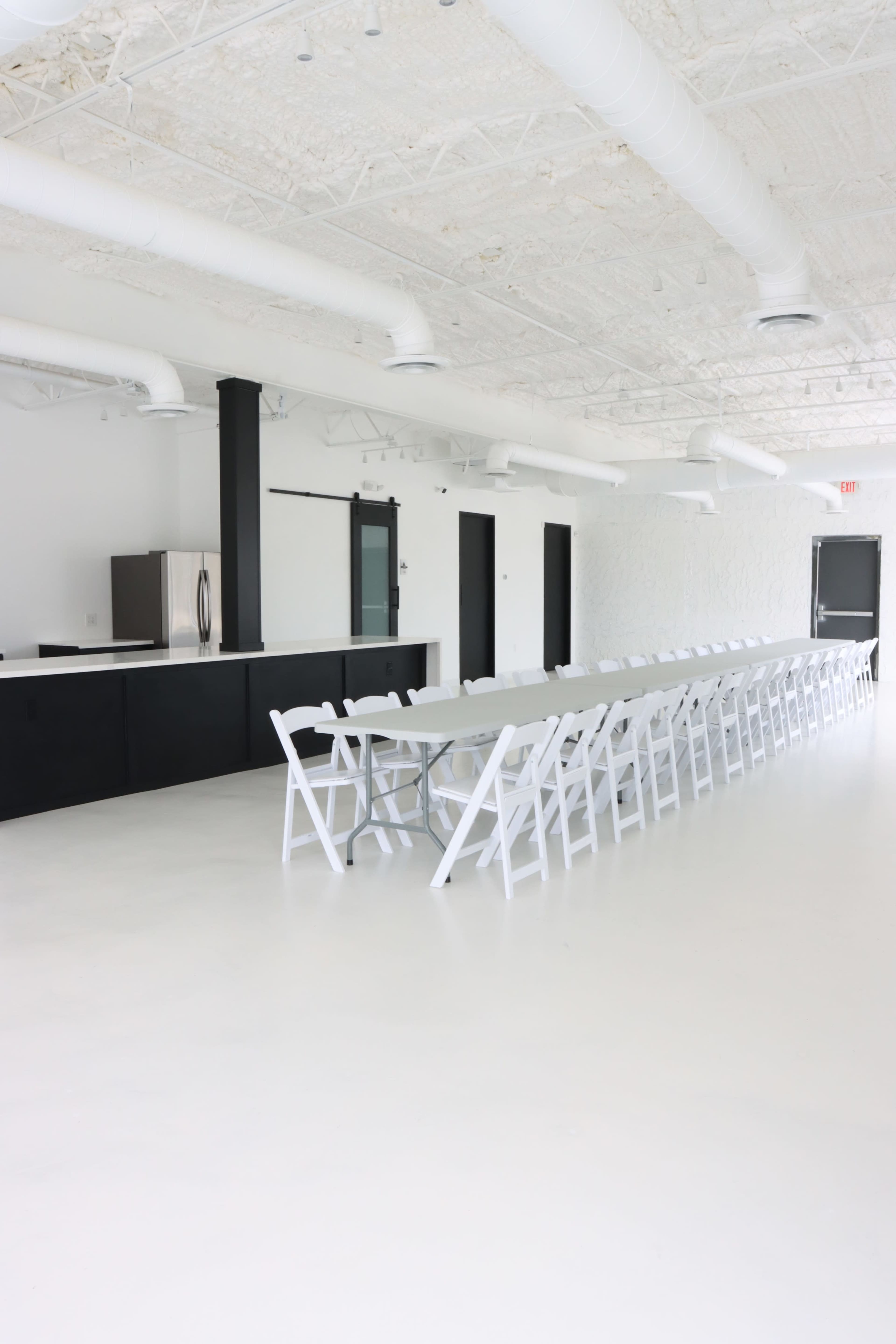 A spacious, minimalist room features a long, white table lined with folding chairs and a black counter against one wall.