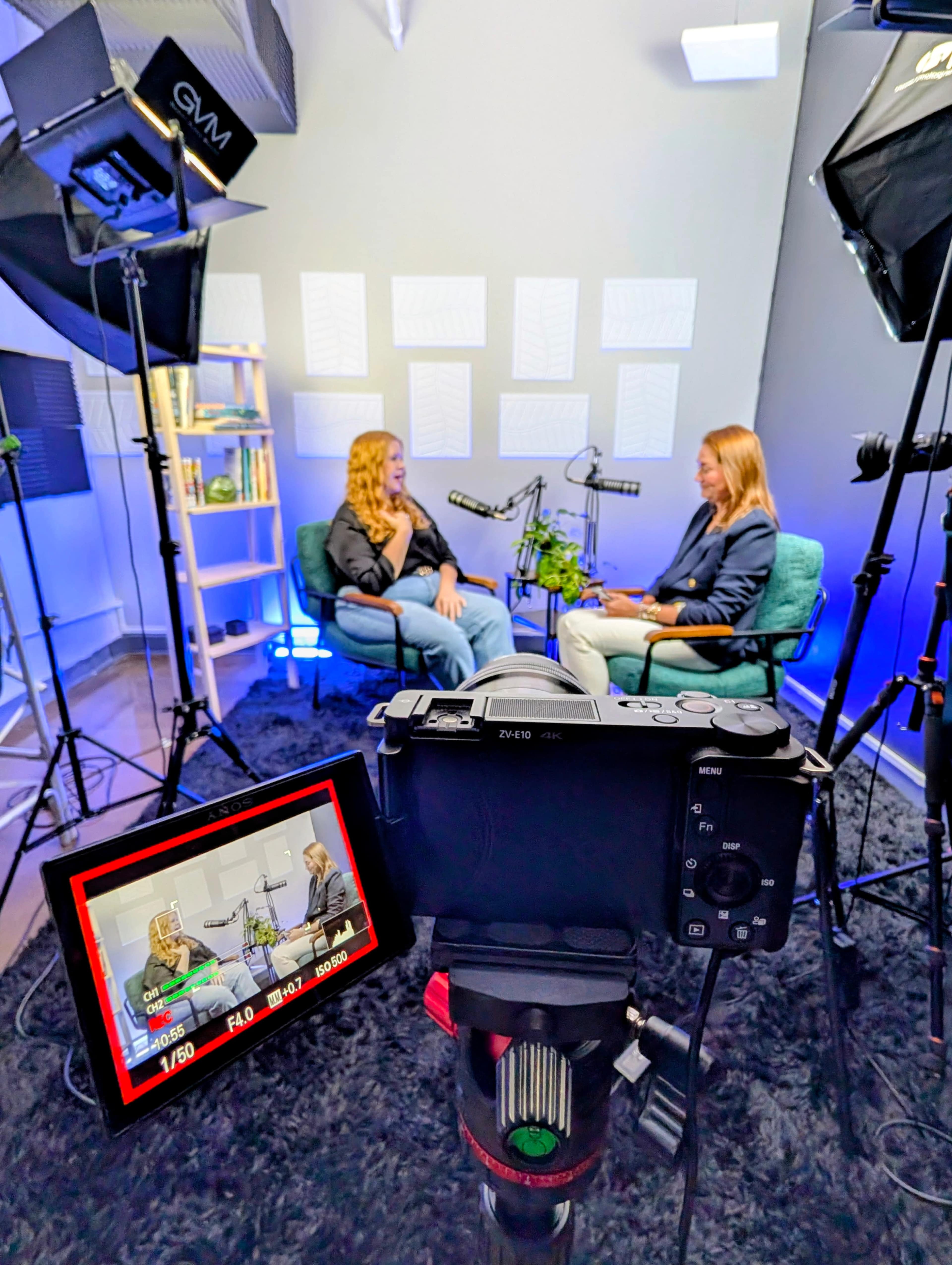A camera captures two women seated in a well-lit studio, engaged in conversation amid professional lighting and backdrop setups.