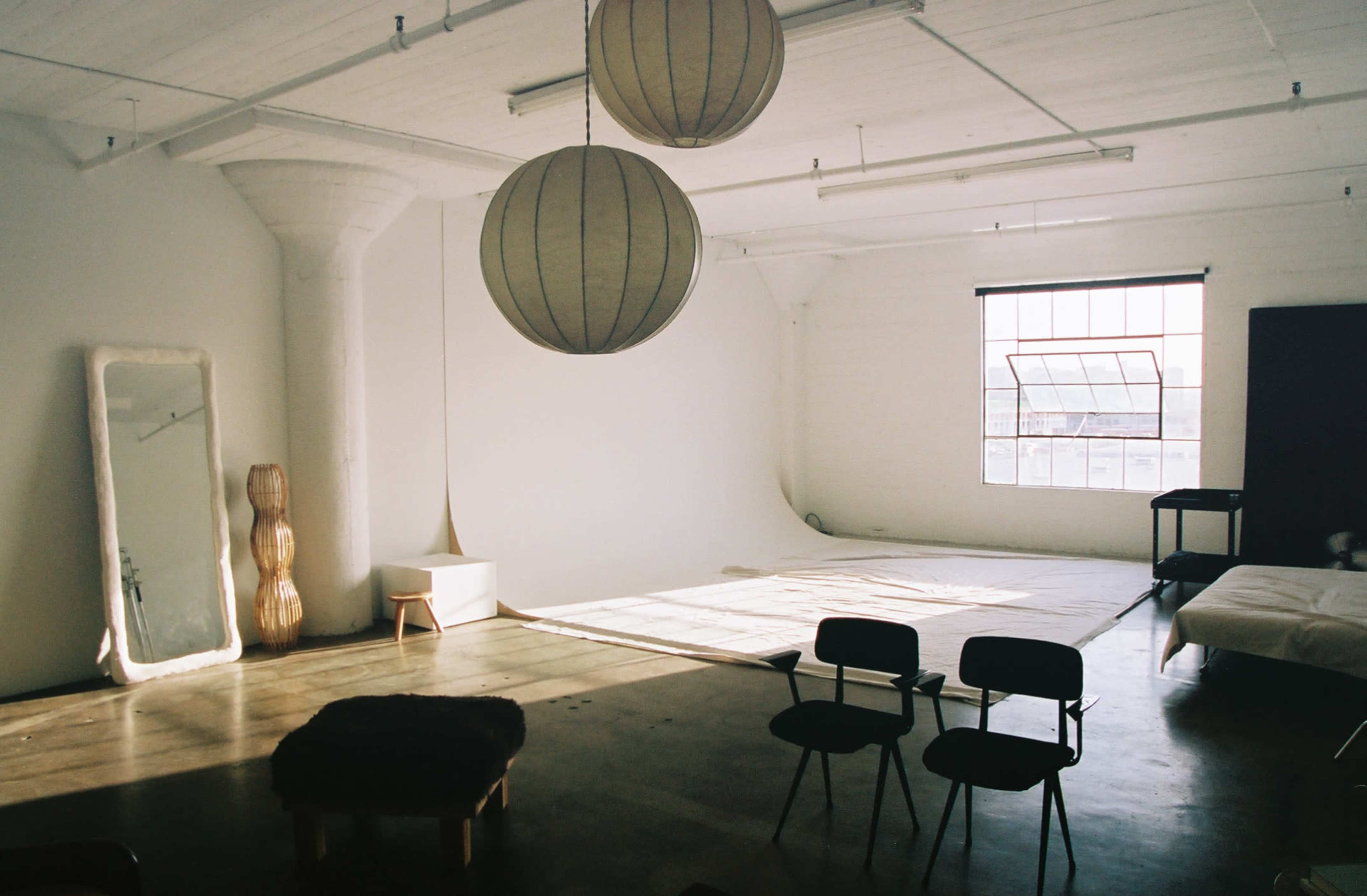 A spacious room with a large window, two spherical pendant lights, and a simple arrangement of furniture including chairs and a full-length mirror.