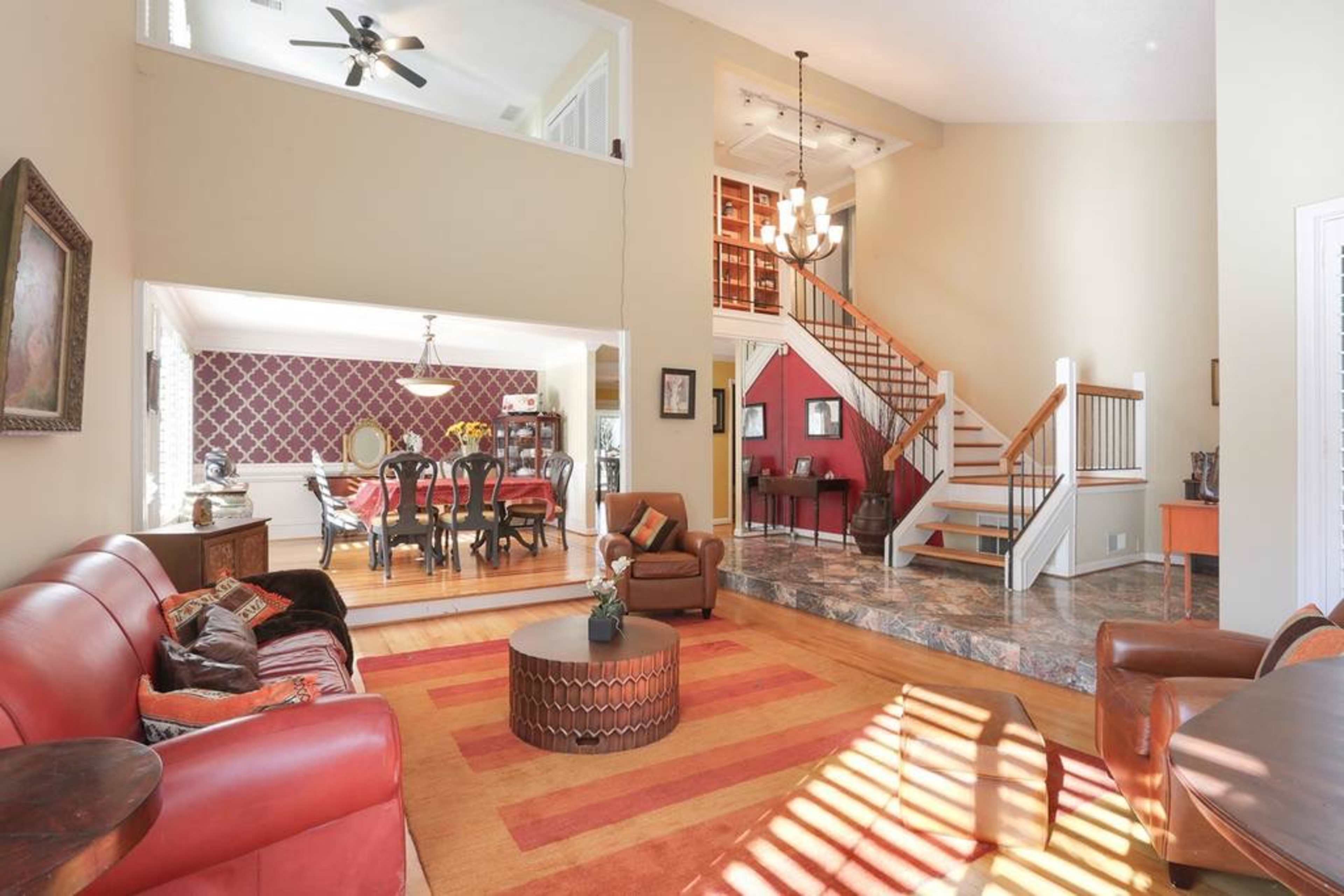 The image shows a spacious living area with brown leather sofas, a round coffee table, and a view of a staircase leading to an upper level and a dining area in the background.
