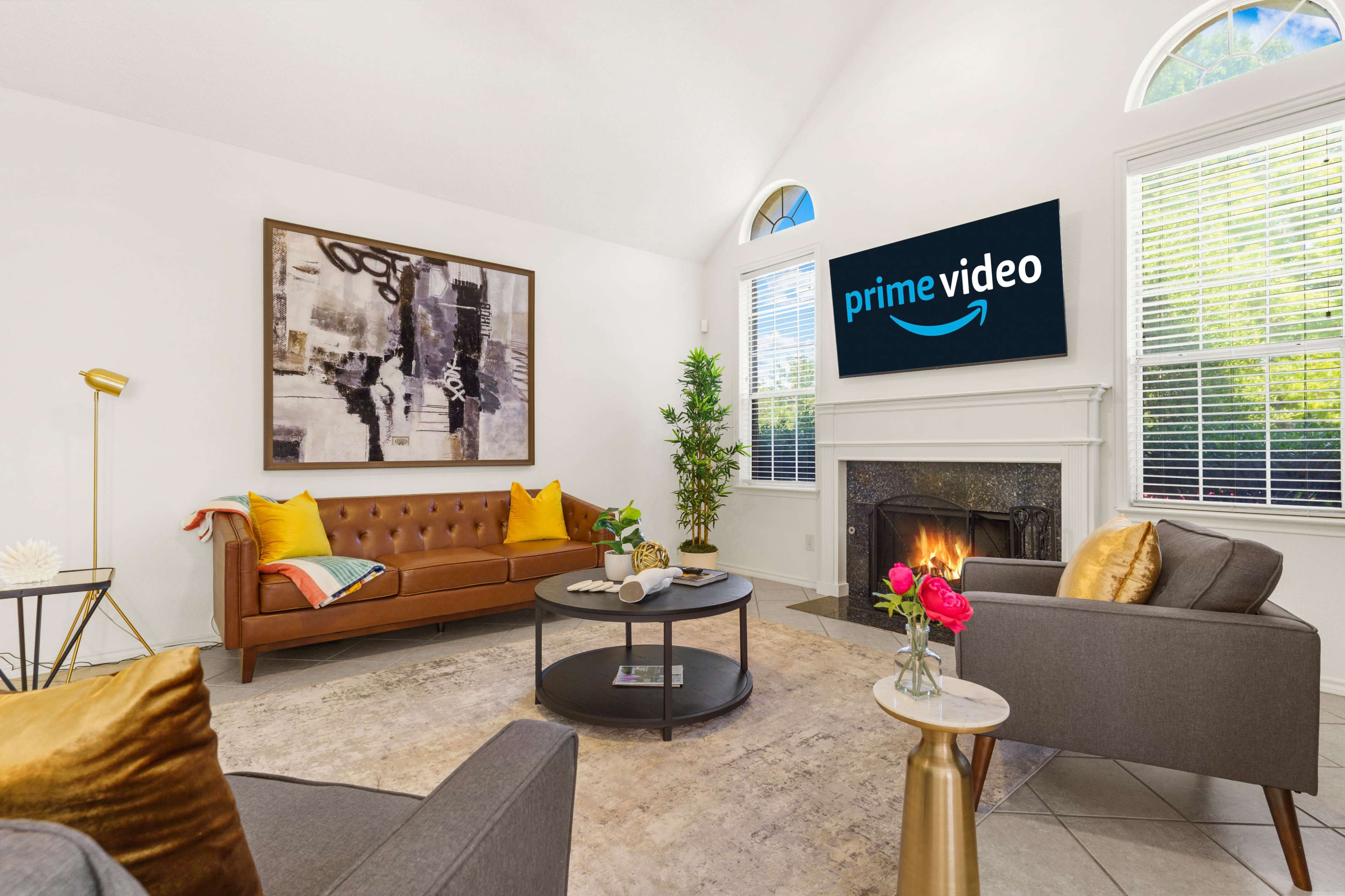 A modern living room featuring a brown leather sofa, a gray armchair, a coffee table, and a television displaying the Prime Video logo above a fireplace.