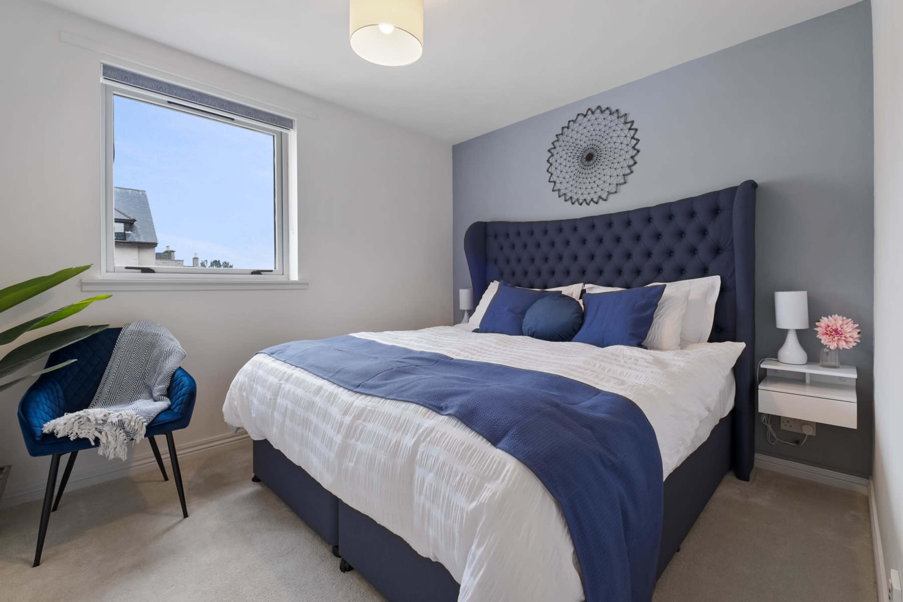 The bedroom features a large bed with blue and white bedding, a tufted headboard, a decorative wall hanging, a chair, and a window allowing natural light in.