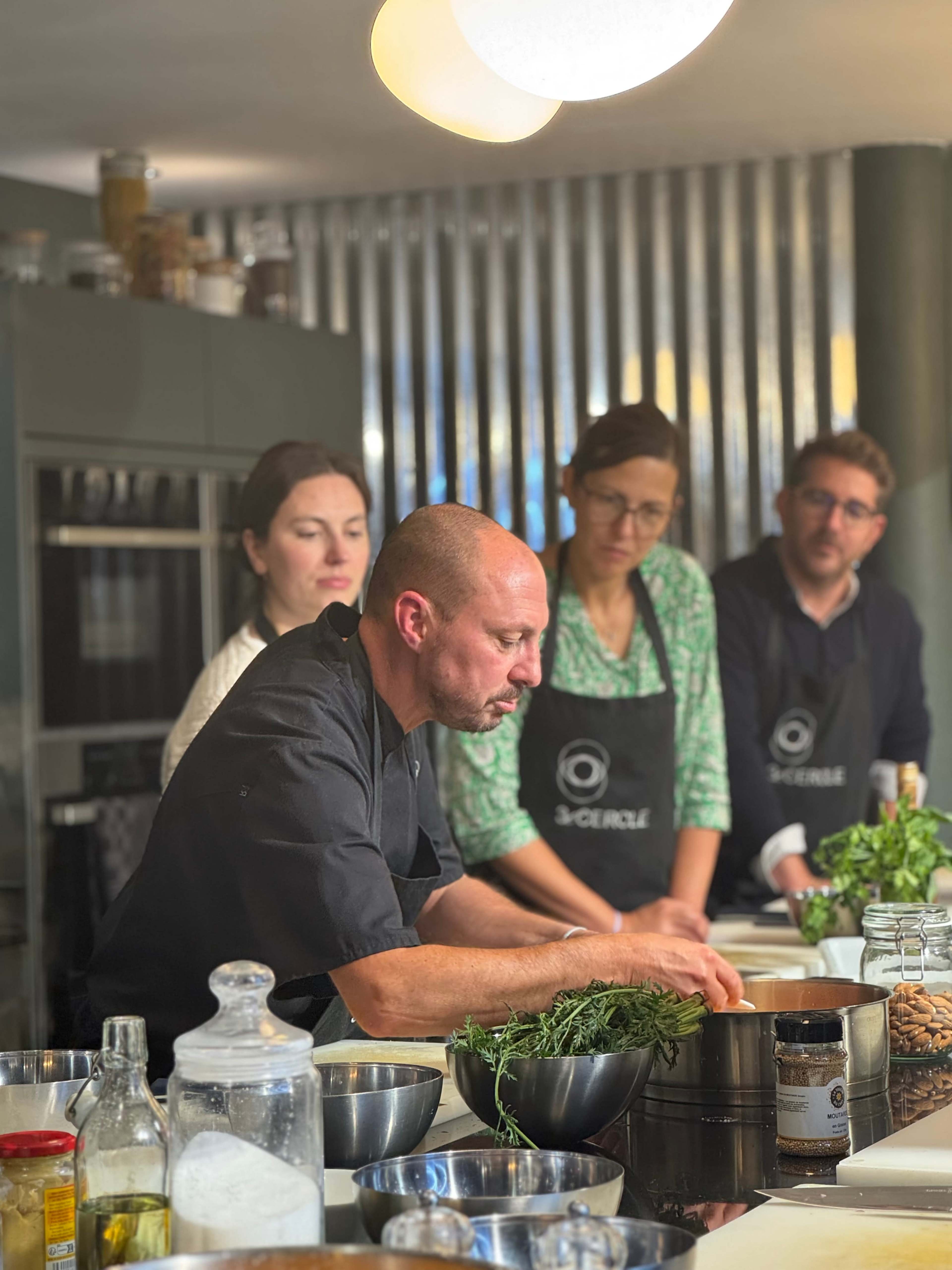 A chef prepares ingredients in a modern kitchen while three observers watch attentively.